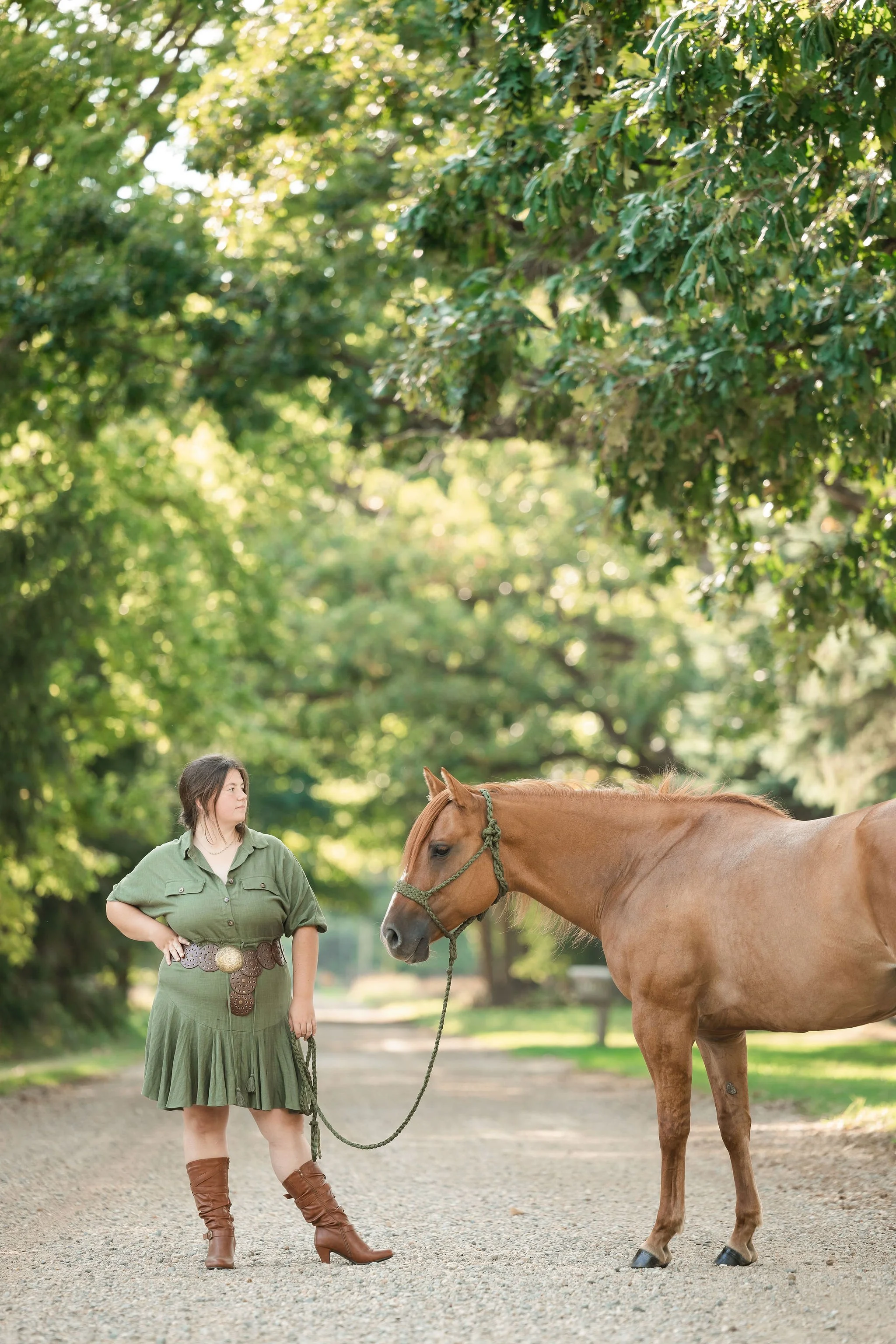 A woman in a green dress and brown boots standing on a gravel path holding a lead rope attached to a brown horse, with trees and sunlight in the background.