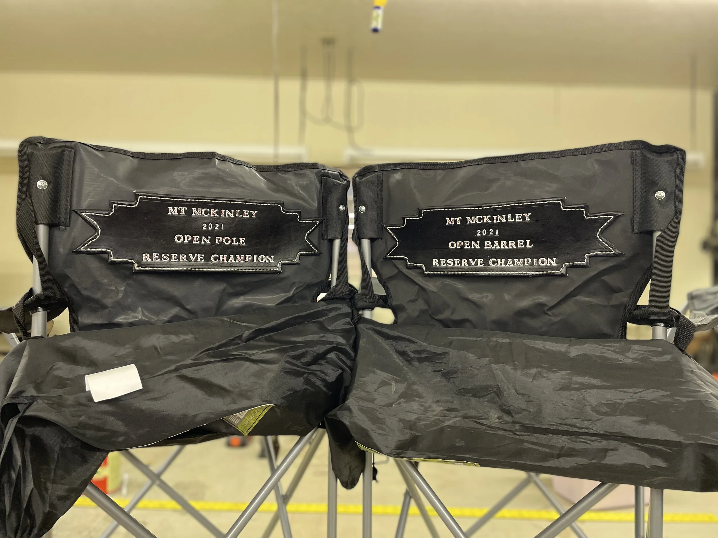 Two black chairs with patches that read "MT MCKINLEY 2021 OPEN POLE RESERVE CHAMPION" and "MT MCKINLEY 2021 OPEN BARREL RESERVE CHAMPION" are positioned next to each other in an indoor setting, with some objects and a beige wall in the background.