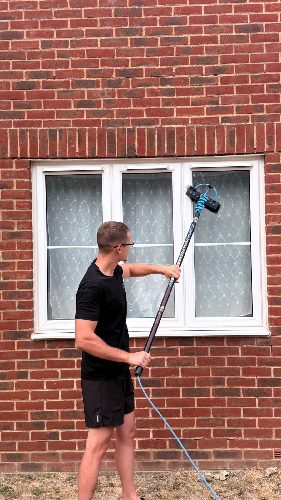 A man in black shorts and t-shirt using a power washer to clean a window of a brick house.