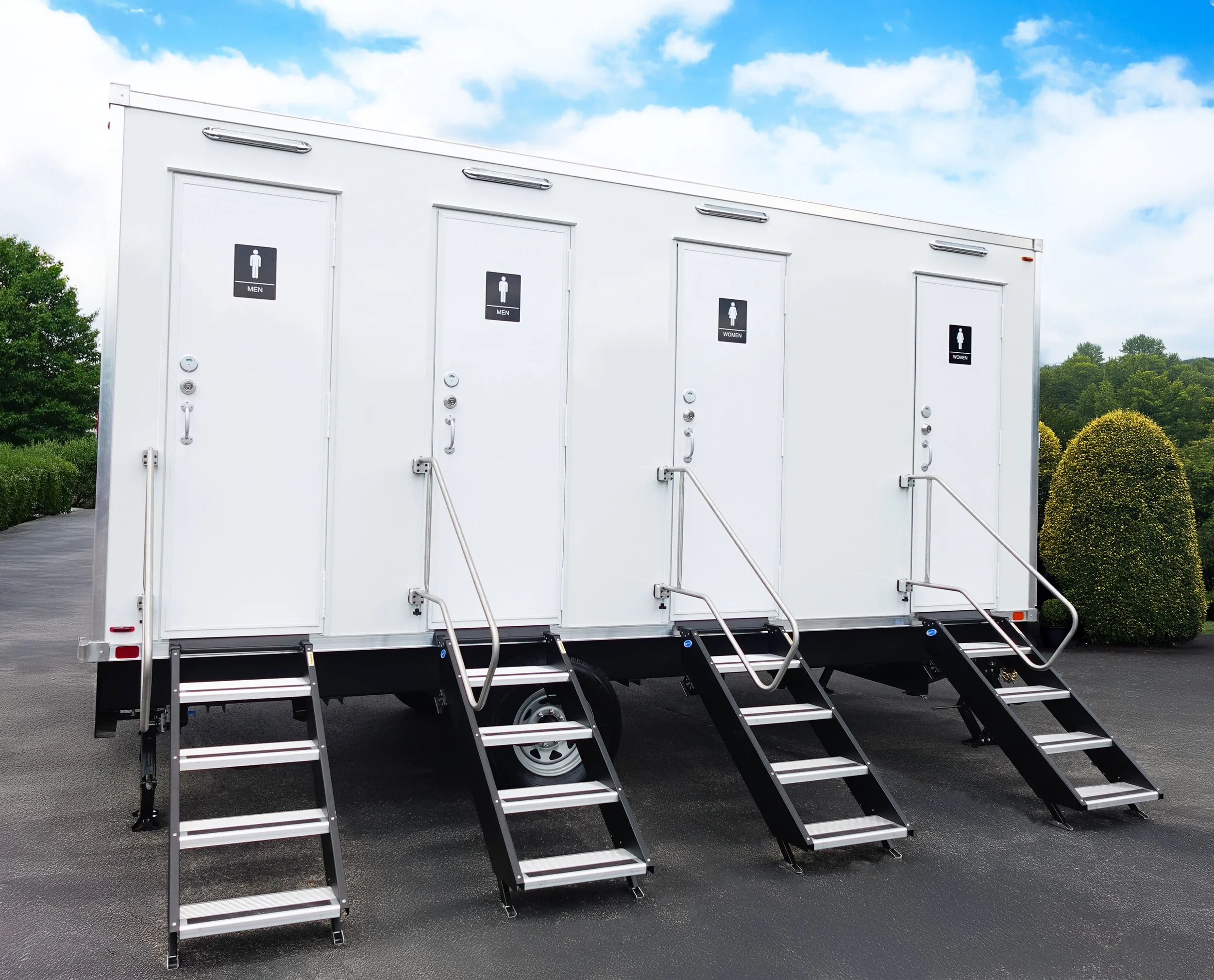 A white portable building with four restroom doors, two labeled for men and two labeled for women, each with stairs and handrails leading up to the entrances, outdoors on asphalt with green trees and shrubs in the background under a partly cloudy sky.