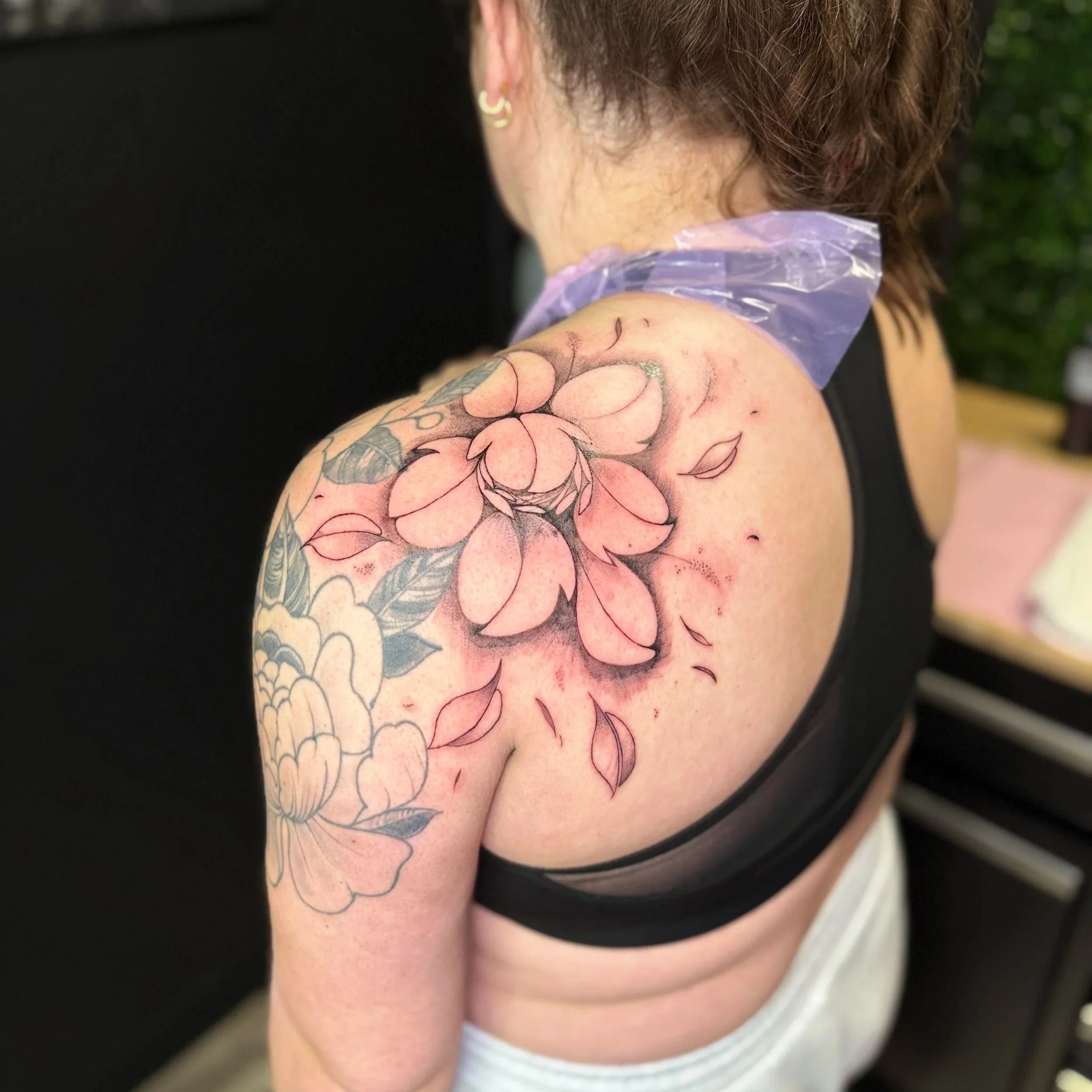 A person with shoulder-length hair receiving a tattoo on their upper arm. The tattoo features roses and leaves in a detailed design.