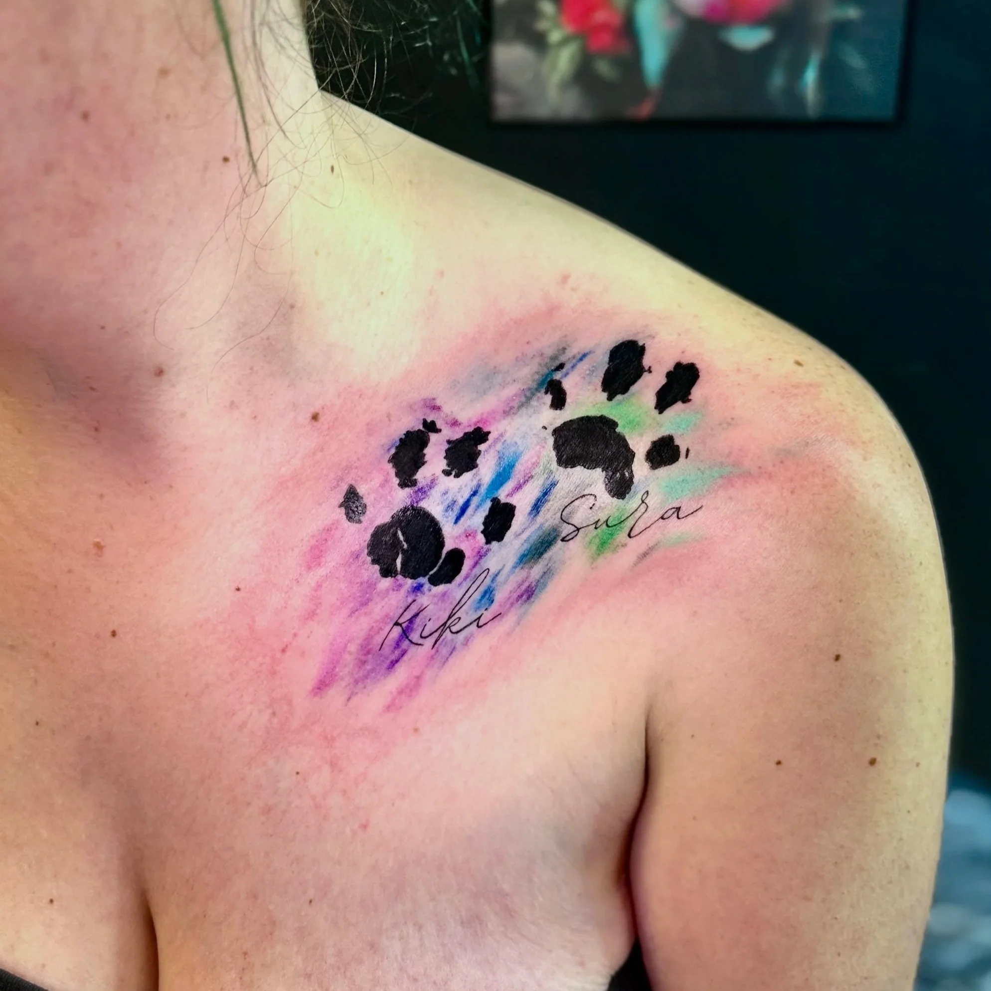 Close-up photo of a person's shoulder and collarbone area with a tattoo of colored dots and scribbles, along with delicate hair and moles on the skin, with a blurred colorful screen in the background.