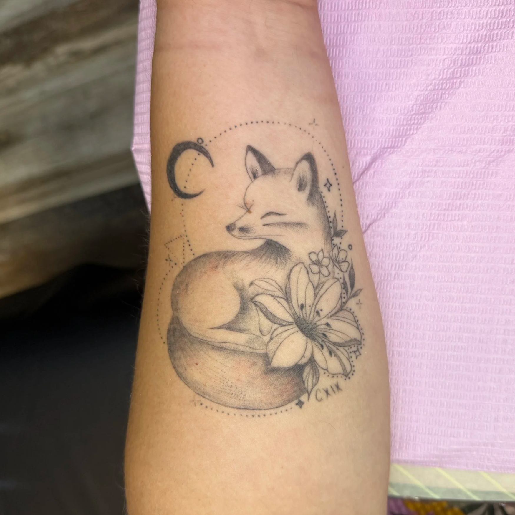 Tattoo of a fox's face surrounded by flowers and a crescent moon on a person's arm.