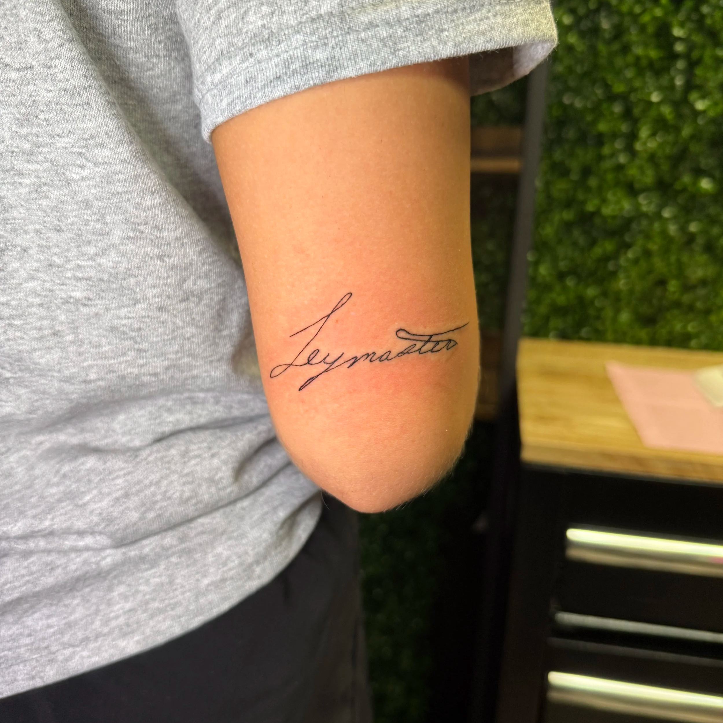 Close-up of a person's arm showing a tattoo with cursive writing that reads 'Leann' or 'Leanna.' The person is wearing a gray shirt, and the background includes green foliage and outdoor furniture.