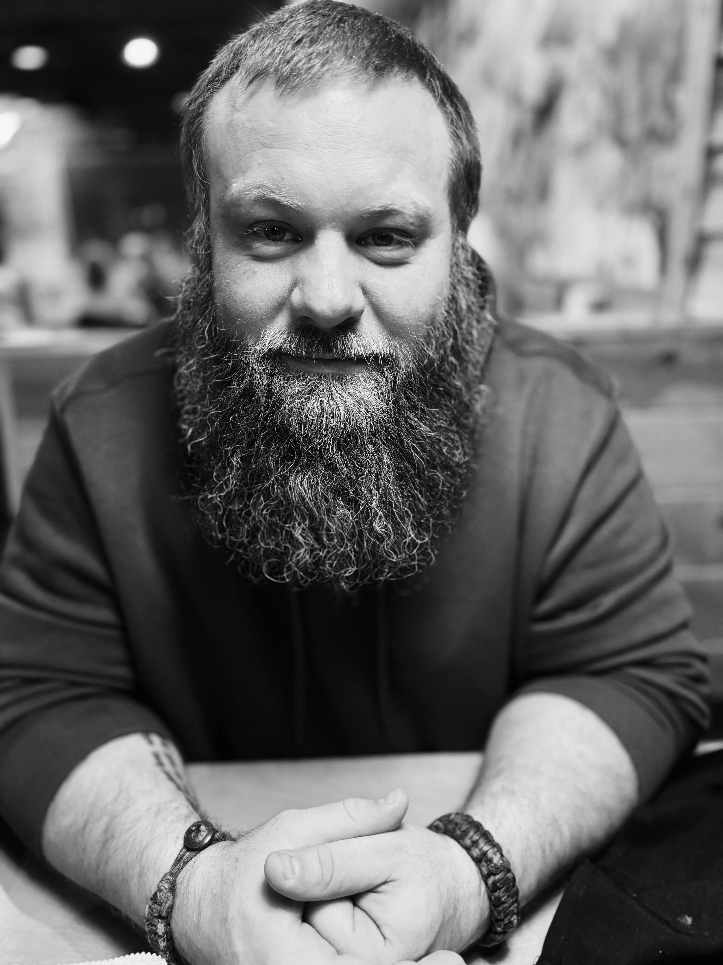 Black and white photo of a man with a beard, looking at the camera, with crossed arms and bracelets on his wrists.