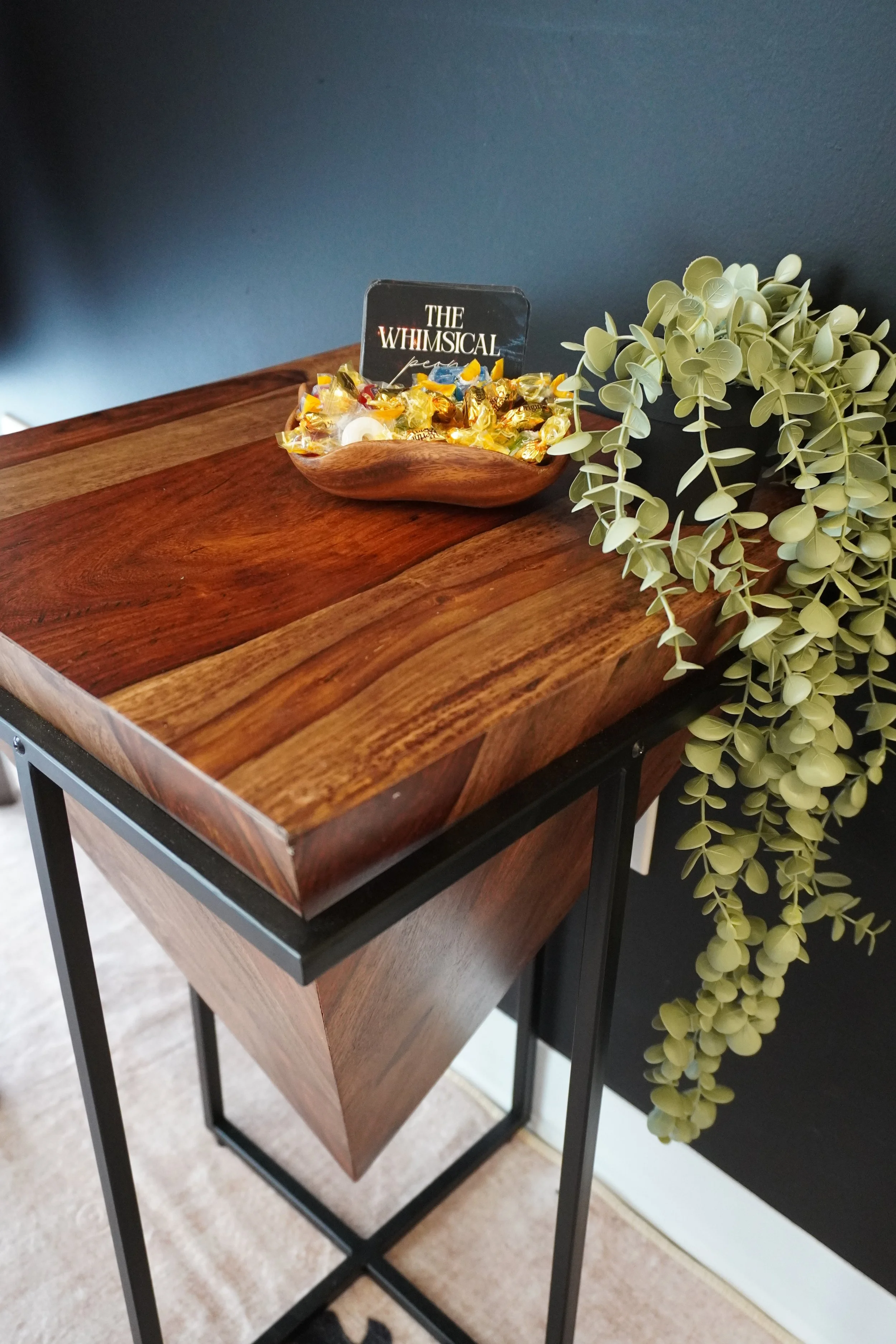 A set of three nested wooden tables with black metal frames, decorated with a black vase of trailing green leaves and a wooden bowl filled with wrapped candies, placed on a dark wall background.