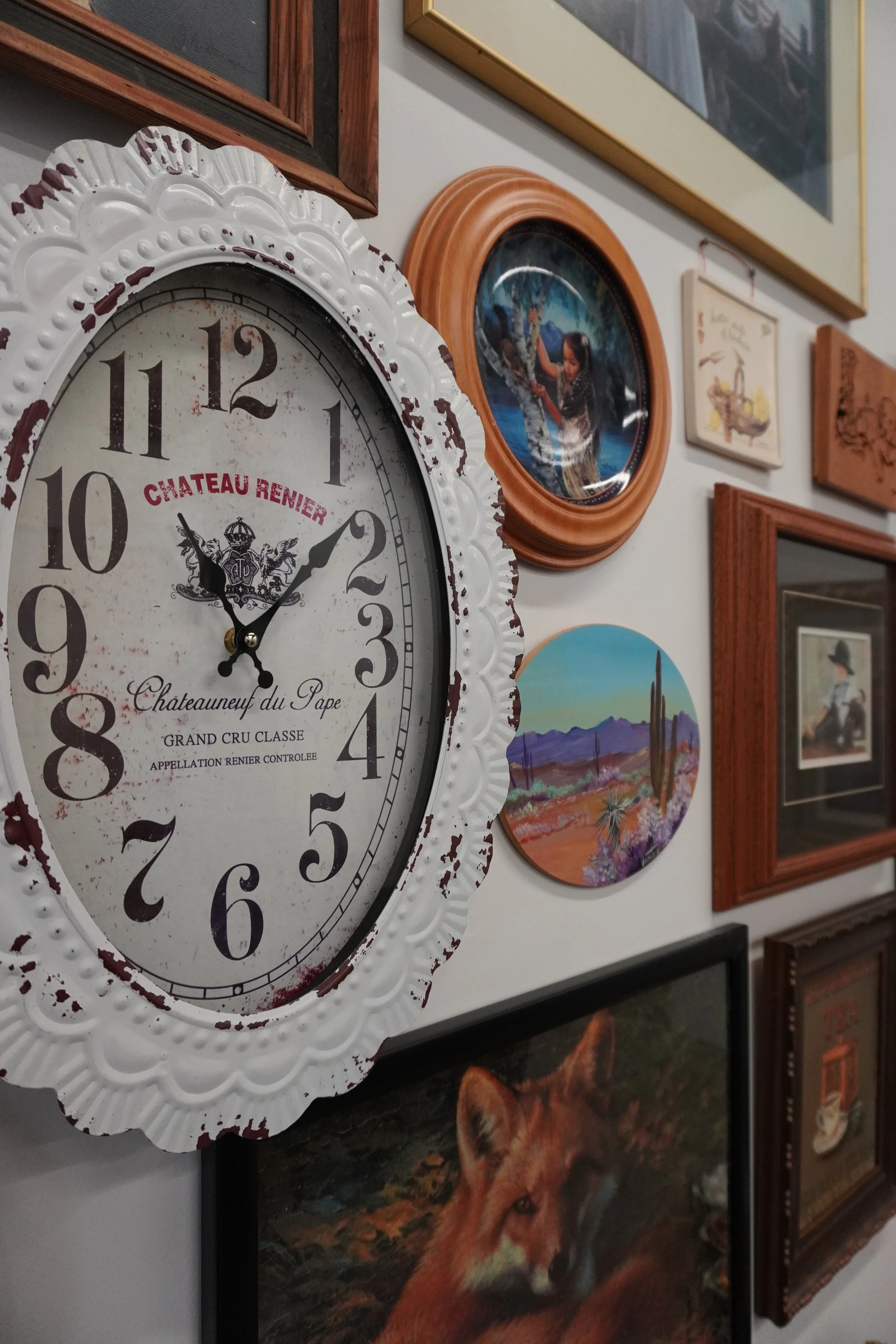 Wall clock with white distressed frame and vintage style face, surrounded by framed artworks including a colorful mountain landscape, a woman crossing a river, and a desert scene with cacti, along with photographs and paintings.