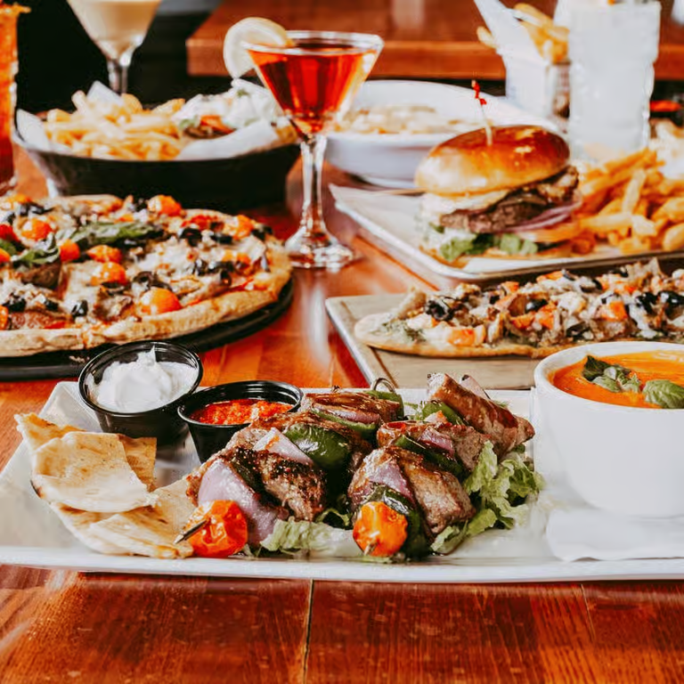 A table filled with various foods including grilled beef and vegetable skewers, pizza with black olives, a bowl of tomato soup, a burger with fries, a pizza, a cocktail, a glass of water, a bowl of salad, and some chips with dipping sauces.