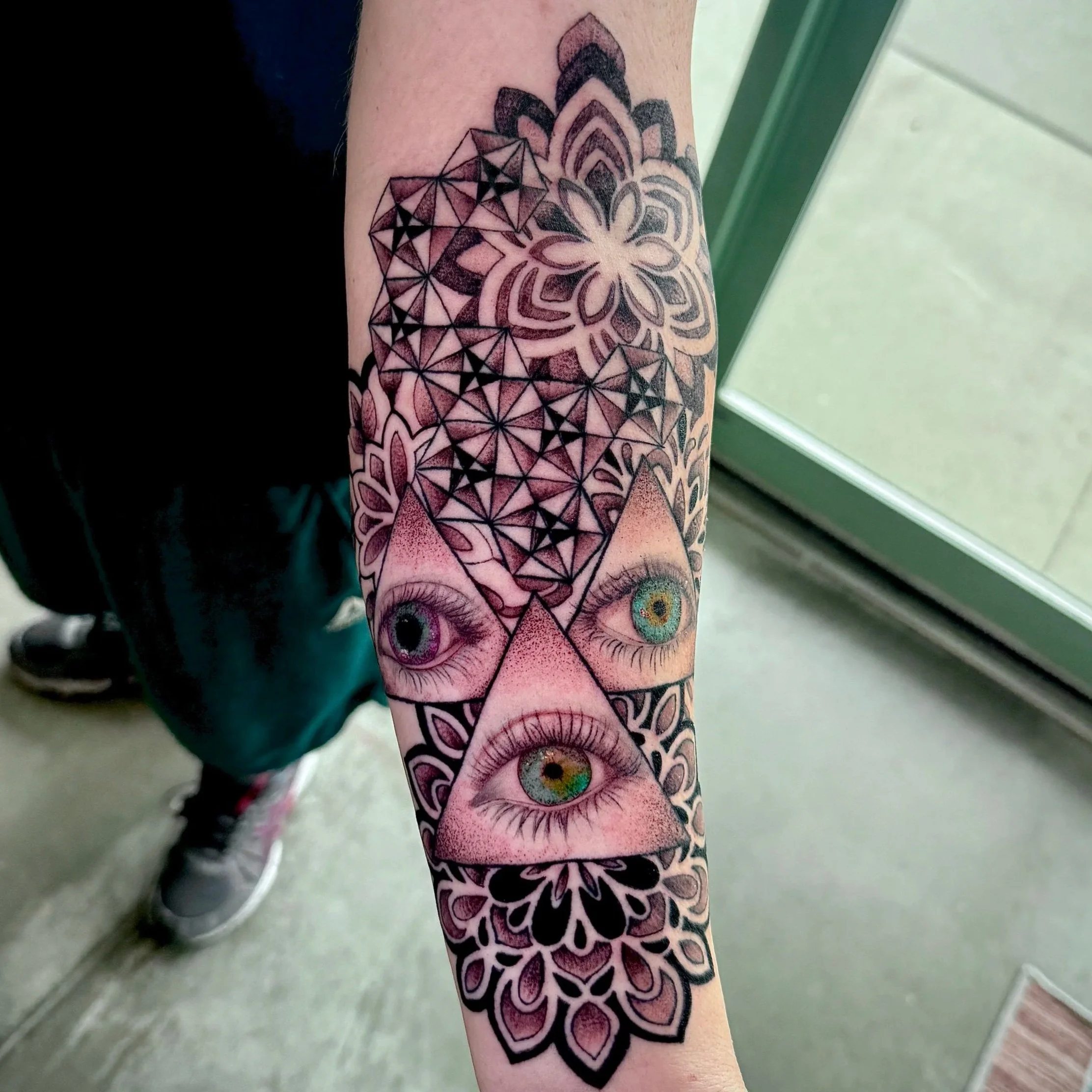 Tattoo of a geometric flower design with a pair of realistic eyes below it on a person's arm.