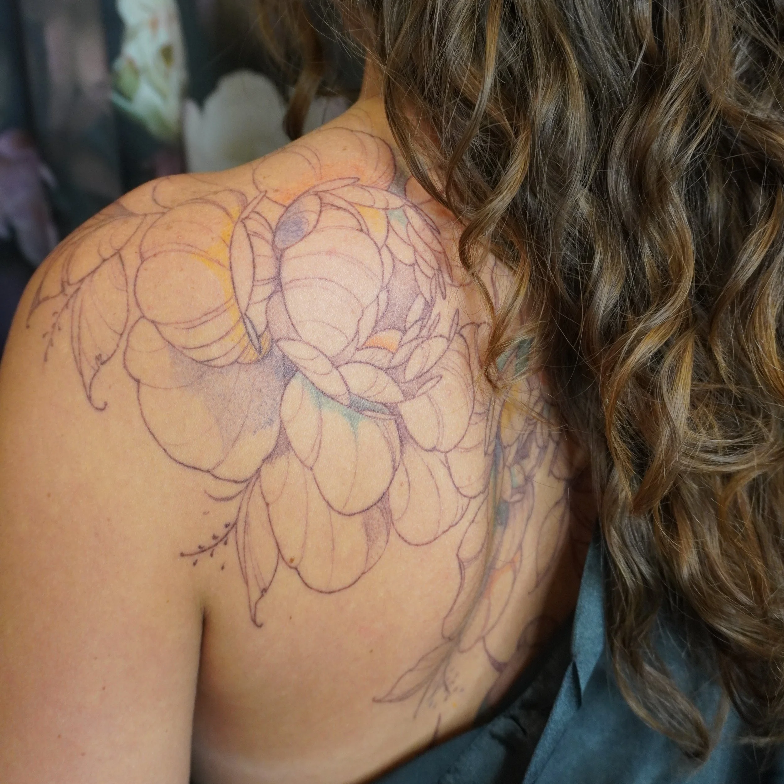 A person's shoulder features a detailed floral tattoo with vivid orange, purple, green, and blue colors, visible on their skin and in their curly, brown hair.