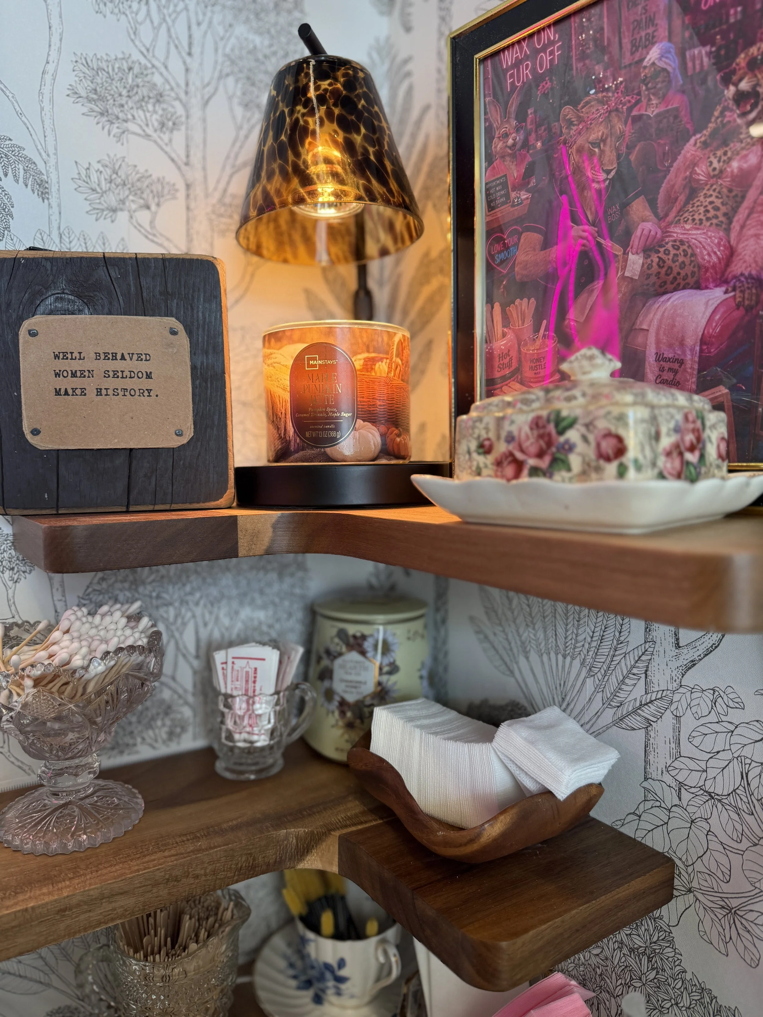 Decorative display with a sign that reads 'Well behaved women seldom make history,' a lit candle in a leopard print lampshade, a framed pink neon sign with various messages, a vintage cake stand with rose patterns holding tissues, a glass cup filled with cotton swabs, and a floral dishcloth, set against a wallpaper with botanical illustrations.
