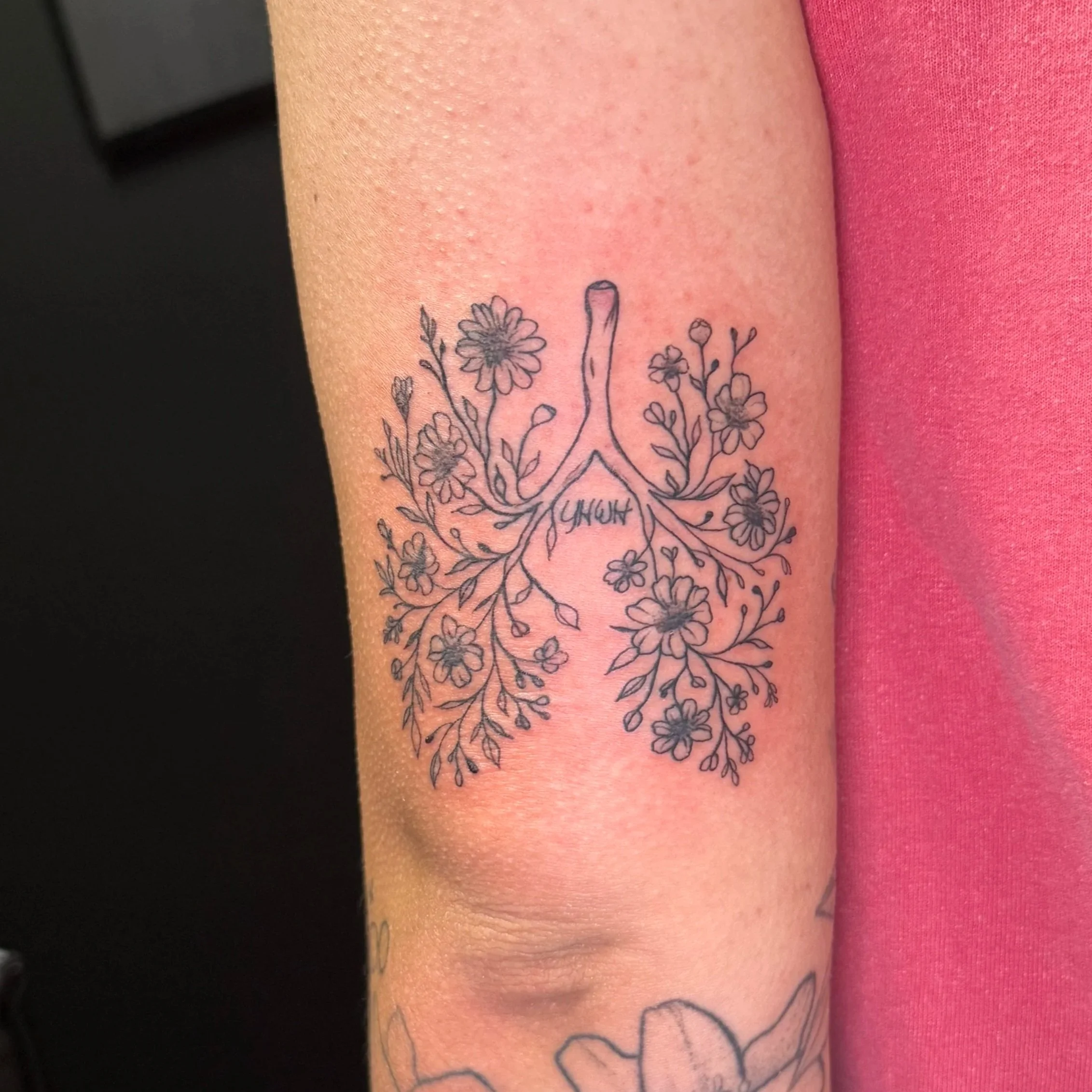 Close-up of a person's arm with a black outline tattoo of flowers and the word 'YATUN' underneath, adjacent to a pink shirt.