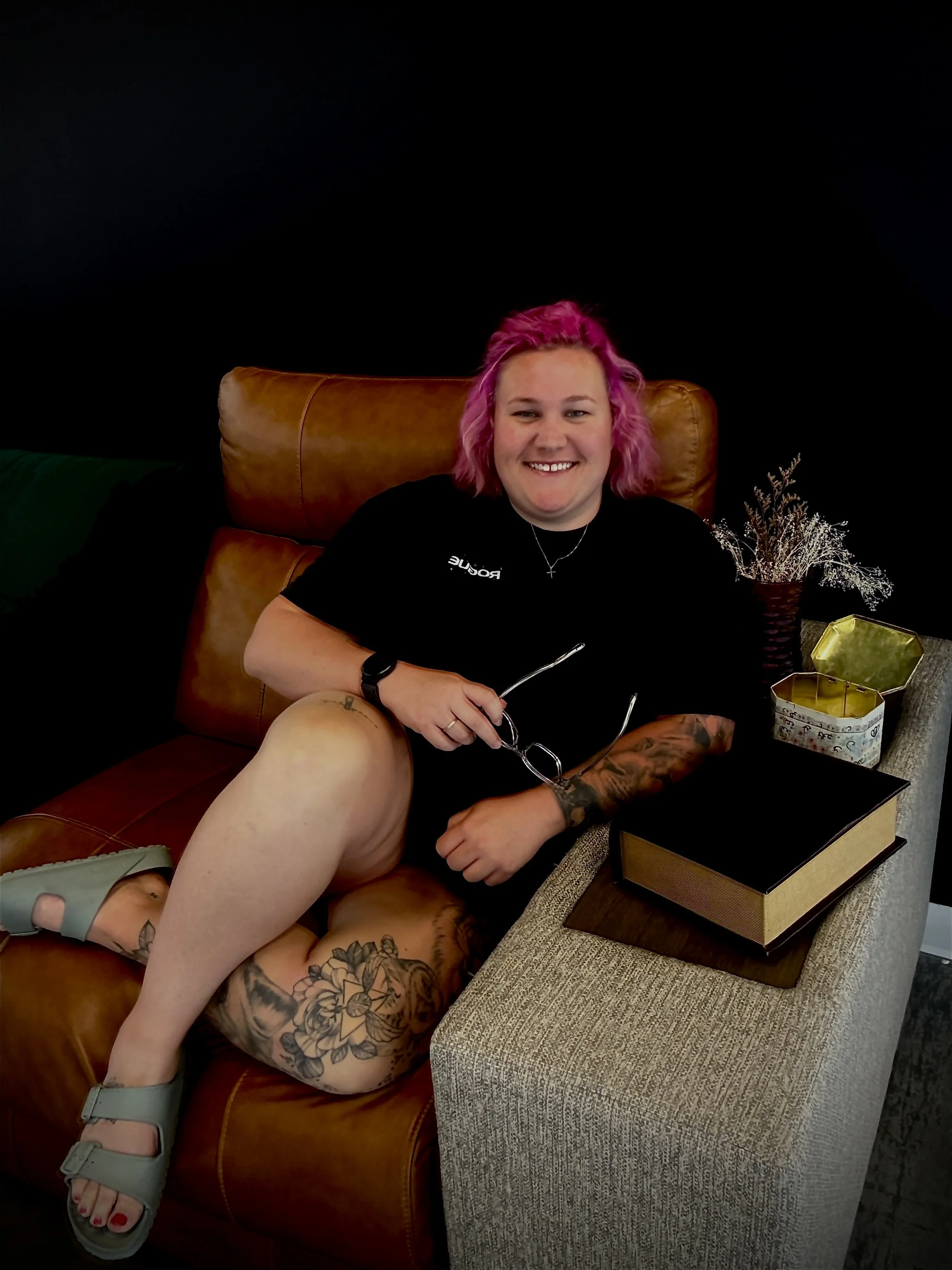 Person with pink hair sitting on a leather chair, smiling, holding glasses, next to a woman lying on her back with tattoos, dressed in black, on a brown couch with her knee resting on the person's lap, in a cozy indoor setting with a dark background and decorative items.