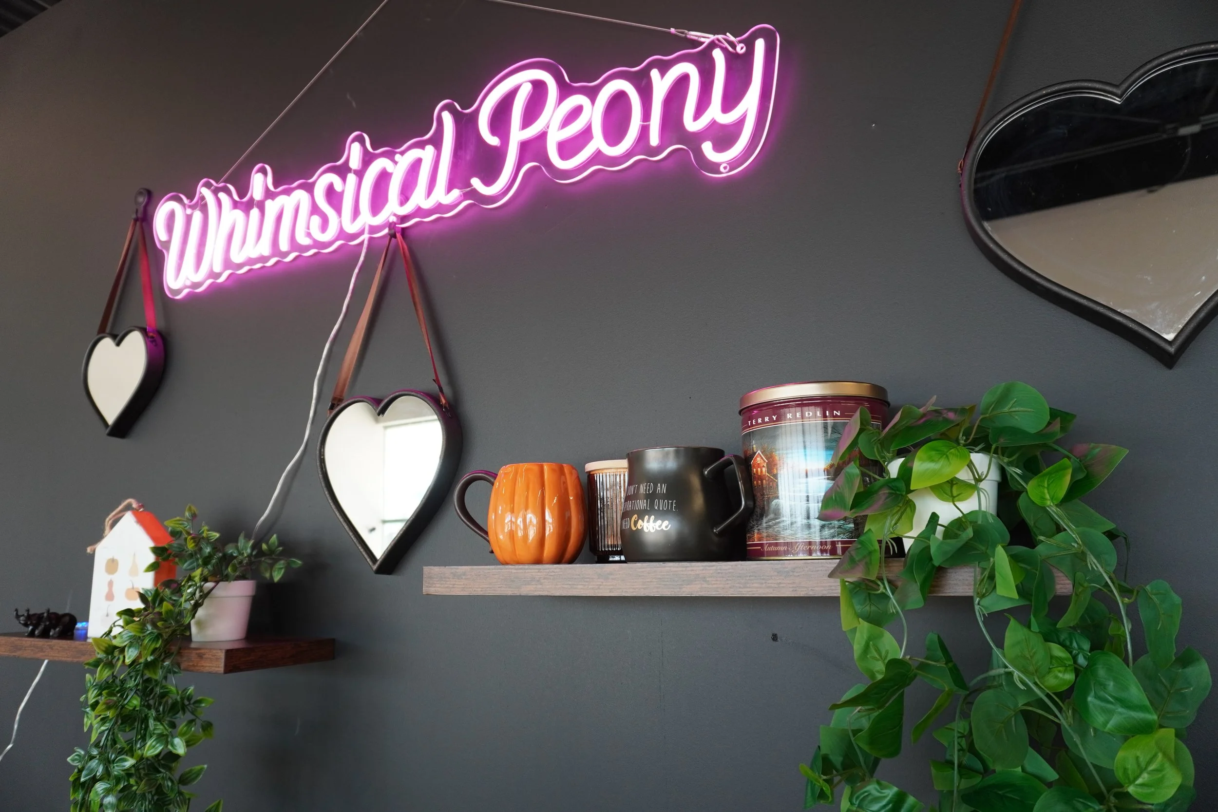 A wall with a pink neon sign that reads 'unusual peony'. There are two heart-shaped mirrors hanging below the sign. On a wooden shelf, there are various mugs and a canister with a nature scene, alongside green plants.
