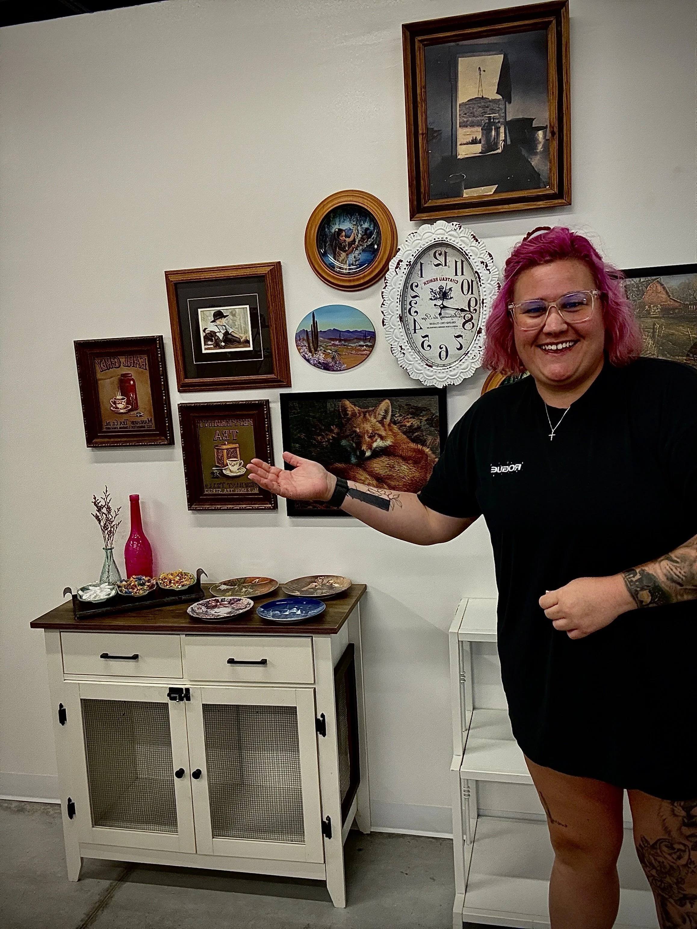 A woman with pink hair and glasses smiling and gesturing towards a white cabinet with framed pictures and a small table with bowls of snacks in front of her, in a room with artwork on the wall.