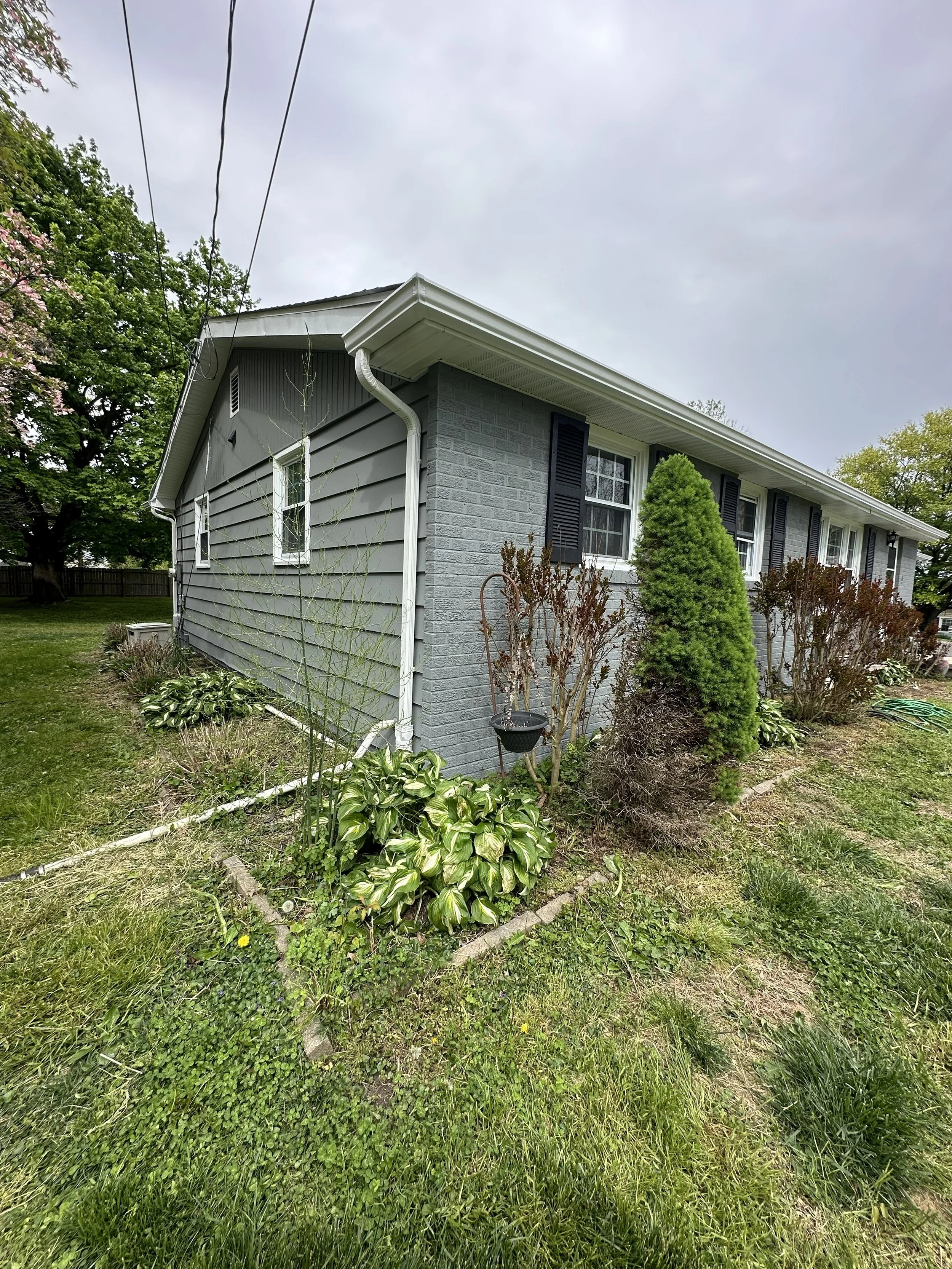 A close-up of the side of a house with gray siding, white trim, and black shutters, surrounded by shrubs and grass, with a cloudy sky overhead.