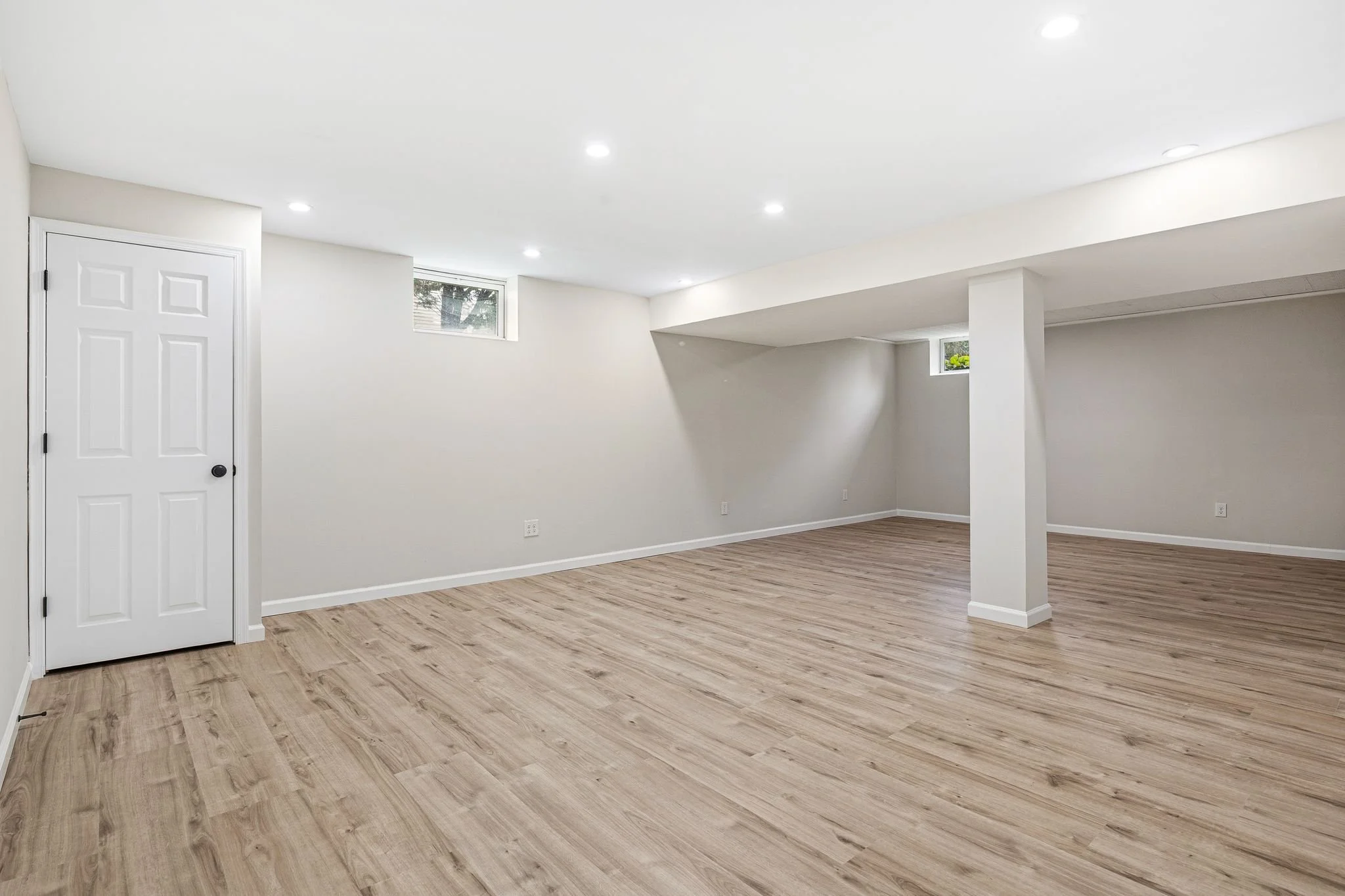Empty room with white walls, wooden flooring, a white door, small high windows, and ceiling lights.