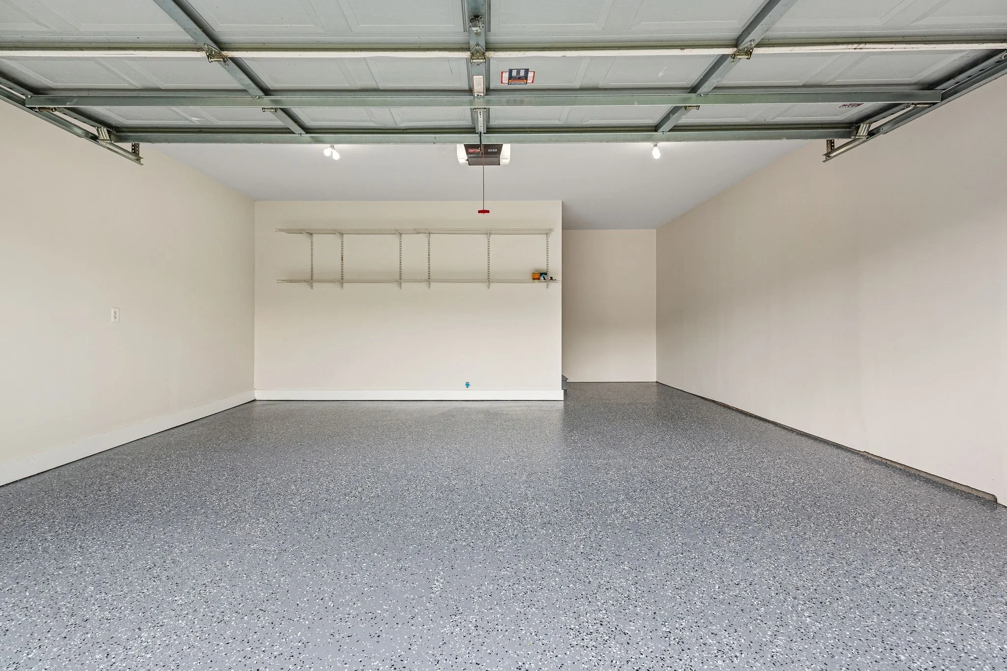 Empty garage with gray speckled epoxy floor, white walls, and a partially open roll-up garage door