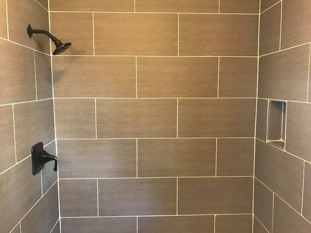 Interior of a walk-in shower with tan wall tiles, a black showerhead, a black faucet, and a small niche for toiletries.