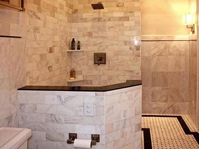 A minimalist bathroom with marble tiles, a small built-in shelf with bottles, a dark countertop, a wall-mounted faucet, and a paper towel holder.