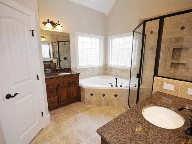 Bathroom with double vanity sink, a corner bathtub beneath two large windows, and a glass shower enclosure. Tiled walls and floor, with a wood cabinet and granite countertop.
