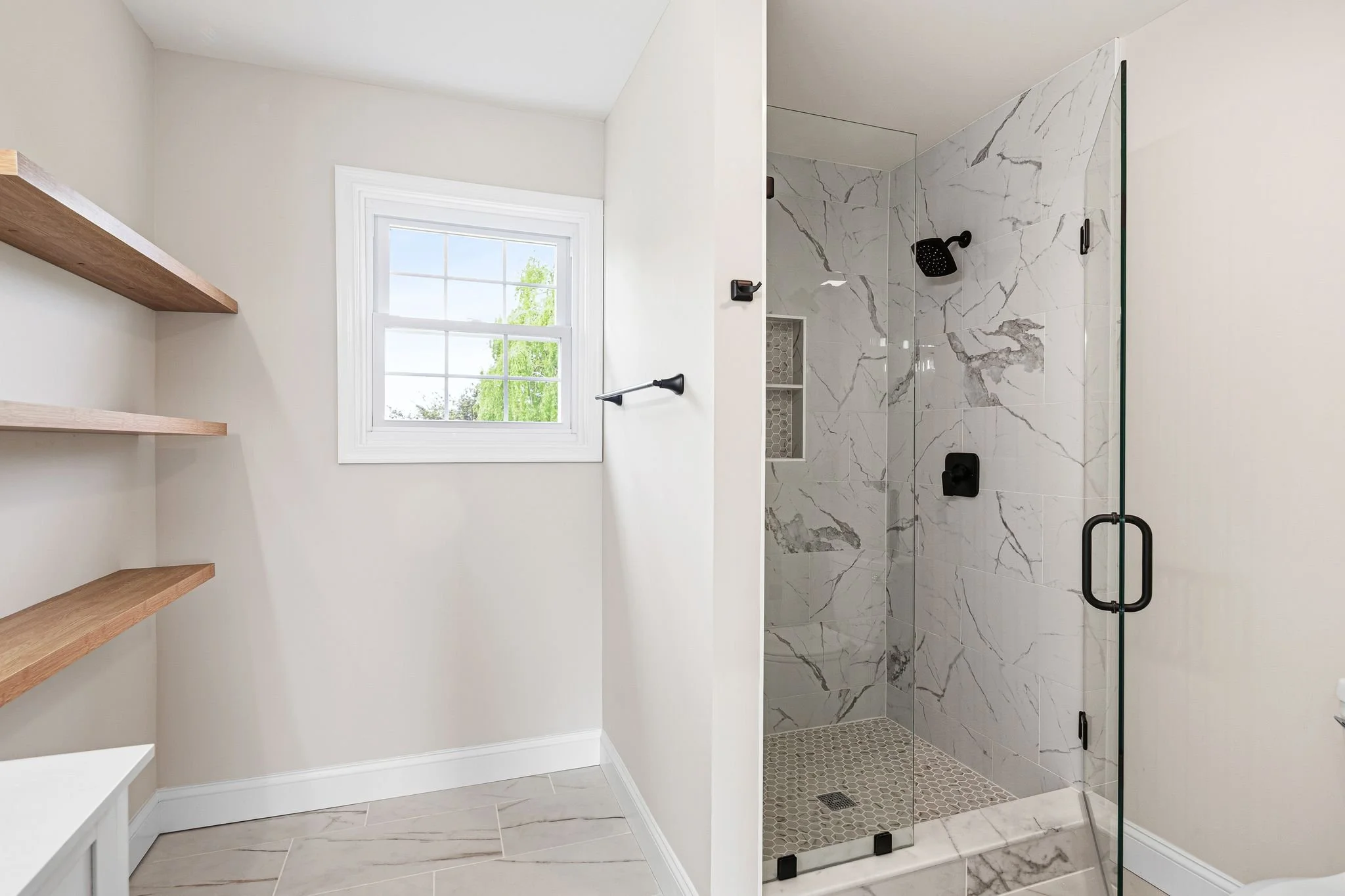 Modern bathroom with a walk-in shower featuring white marble tiles with gray veining, a glass door with black hardware, a small window with white trim, and two wooden shelves on the wall.