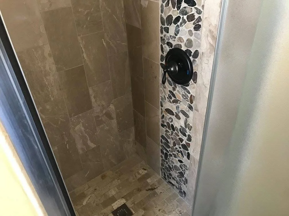 Inside a shower with beige and pebble-patterned tiles, a black faucet handle on the wall.
