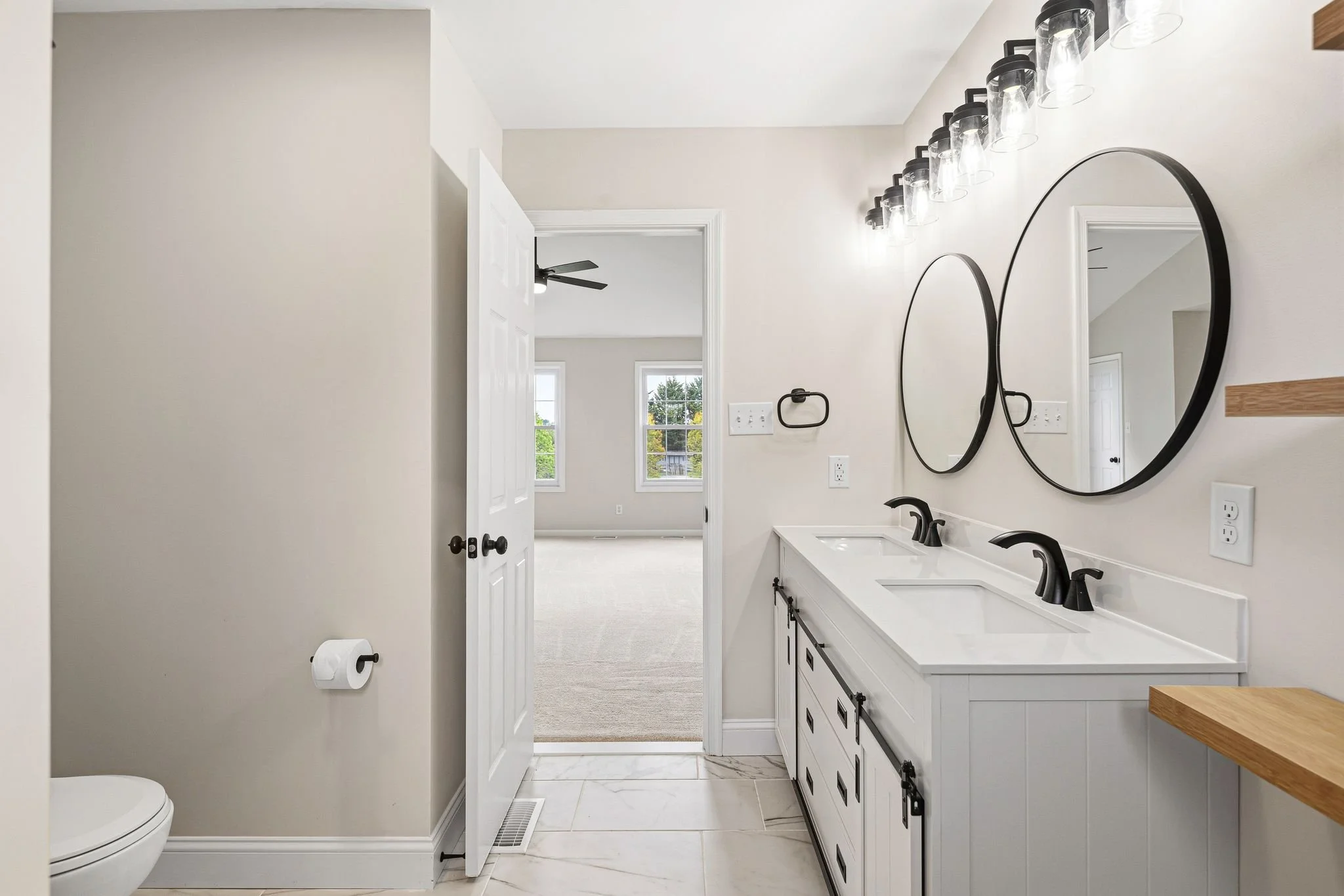 Bathroom with double sinks, oval mirrors, and modern black fixtures, with a view into a bright bedroom through an open door.