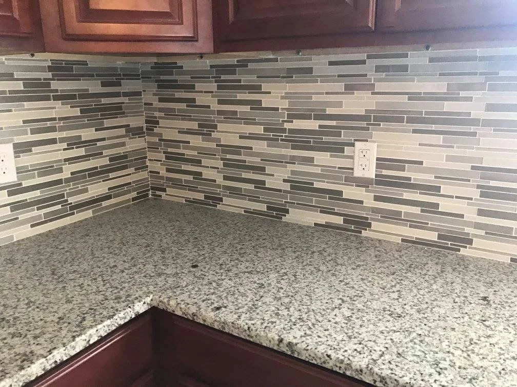 Kitchen countertop with a granite surface, a mosaic tile backsplash in shades of gray and beige, and dark wooden cabinets above.