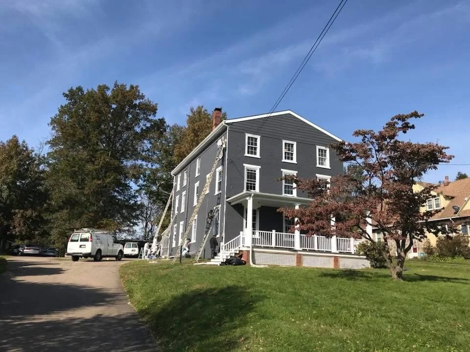 A large gray house with white trim is being painted or repaired with the help of multiple ladders and workers on a sunny day. There are trees and parked vehicles nearby.