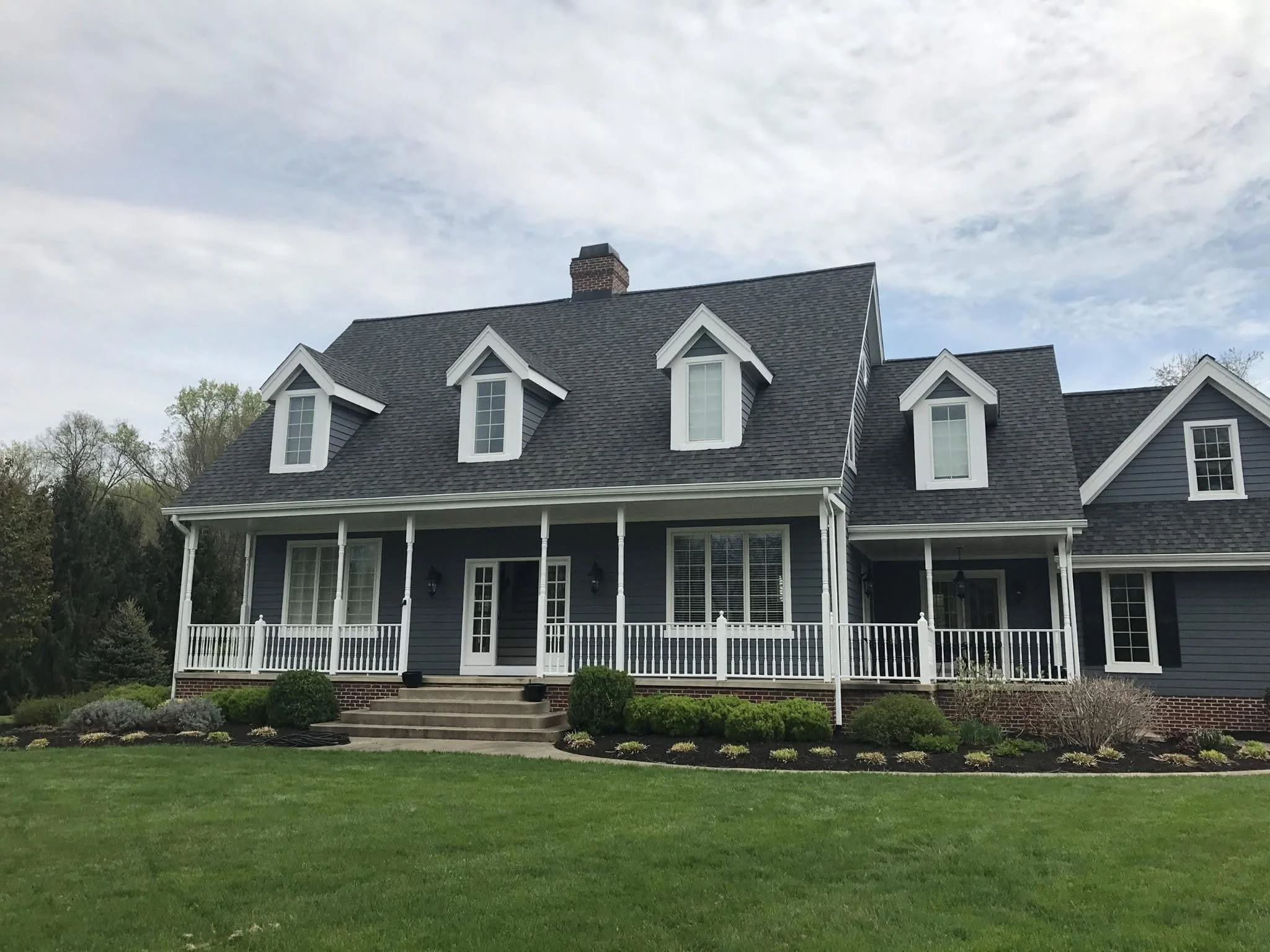 A large two-story house with a dark gray roof, dormer windows, white trim, and a front porch with white railings. The house is surrounded by a well-manicured lawn and garden with shrubs and flowers. The sky above is partly cloudy with some blue visib