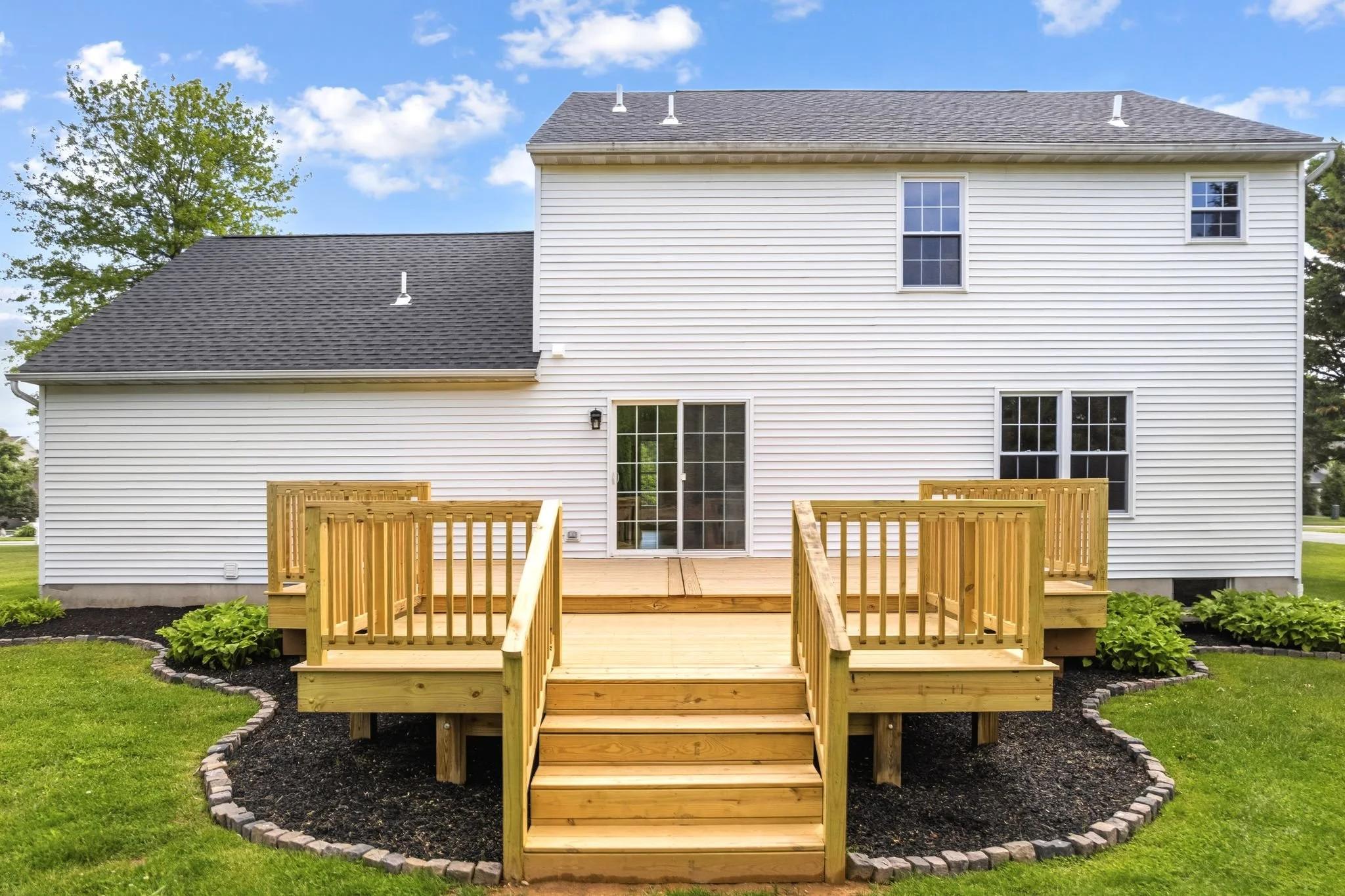 Backyard of a house with a wooden deck and stairs, surrounded by a garden with green grass and plants, and a white house with sliding door and windows, under a blue sky with clouds.