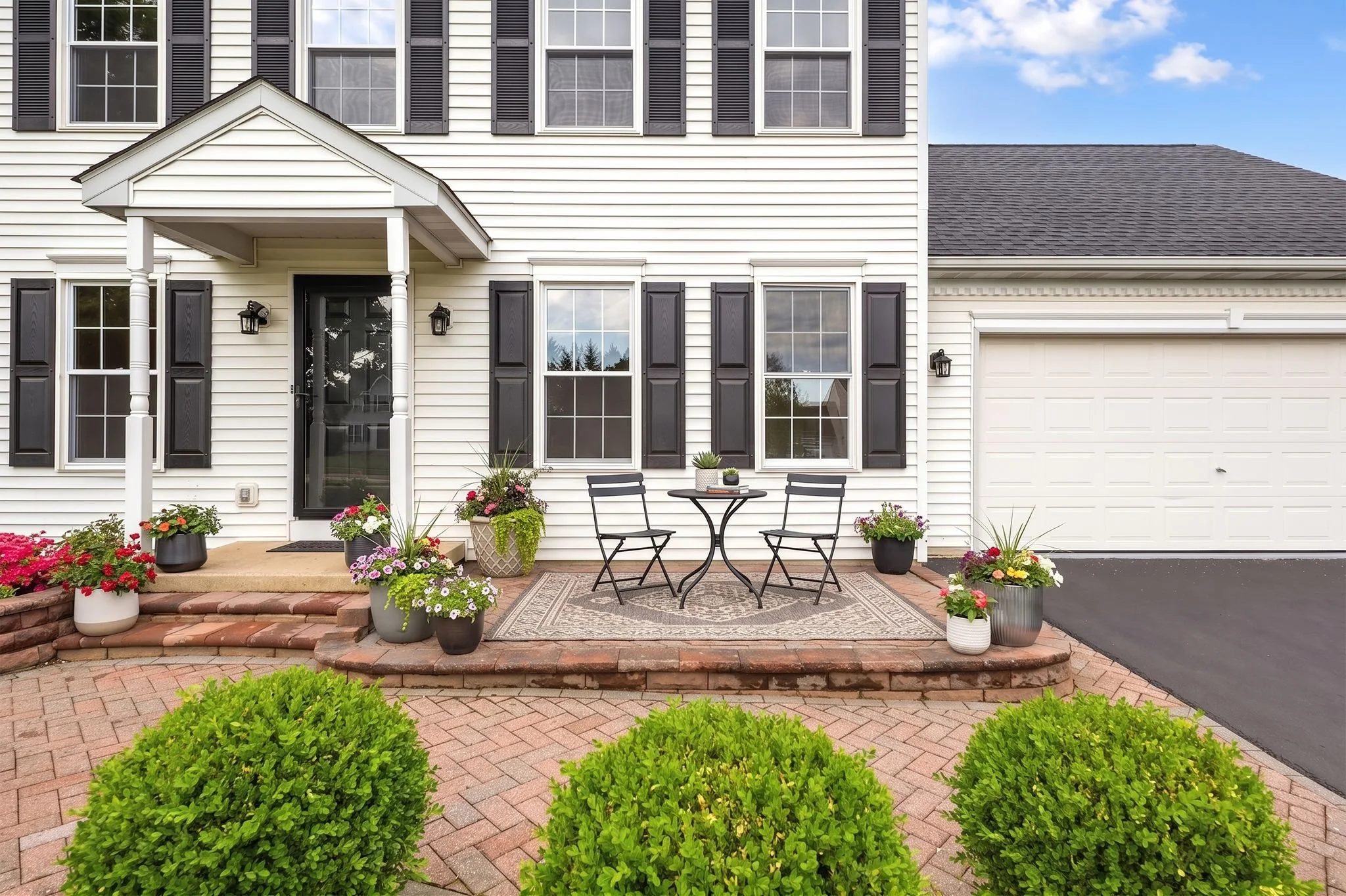 Front of a white house with black shutters, black front door, porch with small table and chairs, and potted flowers. Bricked pathway leading to the porch, green bushes in the foreground, and a partly cloudy blue sky.