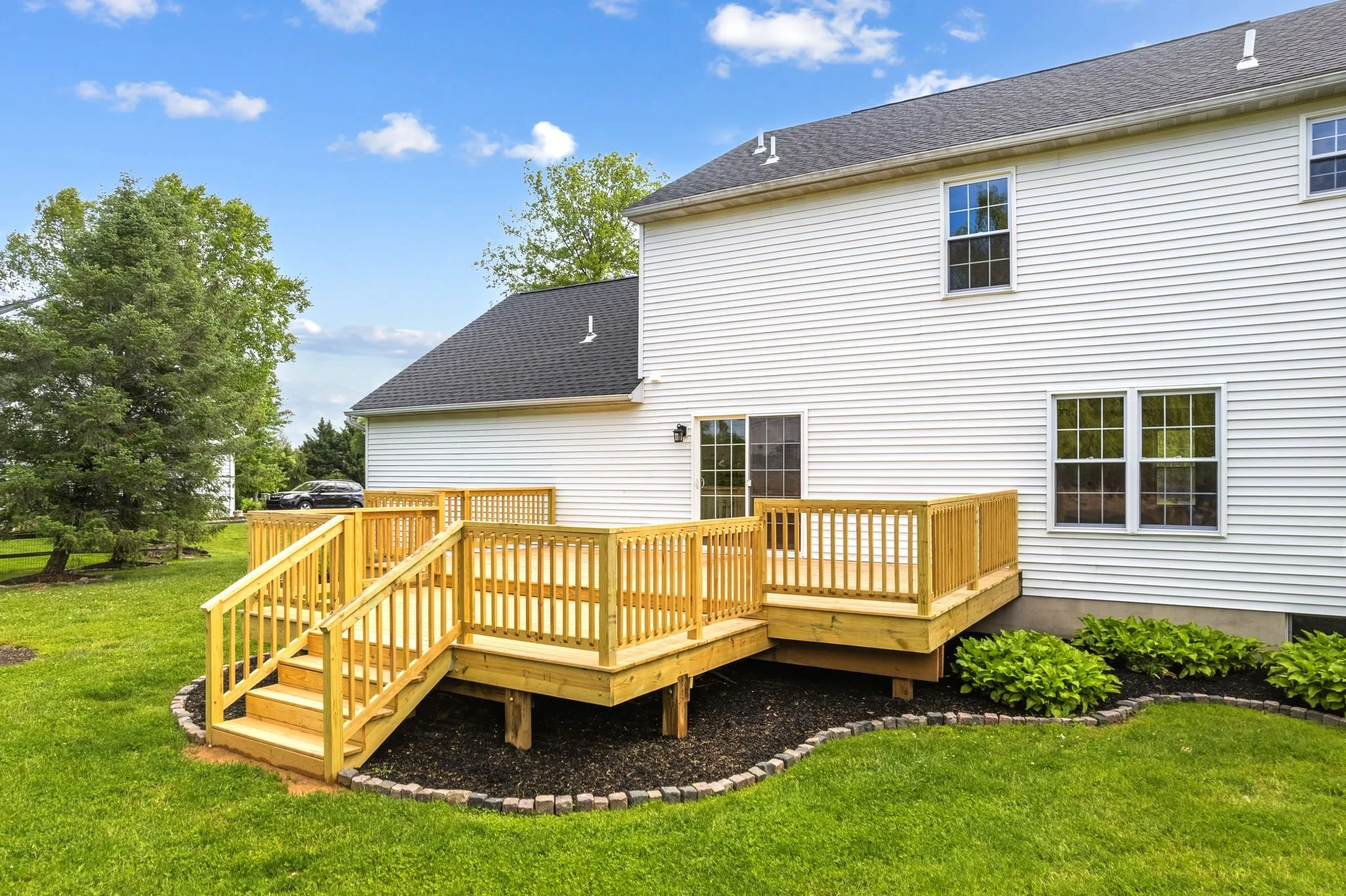Backyard with a new wooden deck attached to a white house with multiple windows, surrounded by green grass, trees, and a blue sky with some clouds.