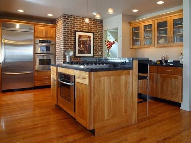 Modern kitchen with wooden cabinets, stainless steel refrigerator, brick accent wall, and an island with black countertop, decorated with a framed picture and flowers.