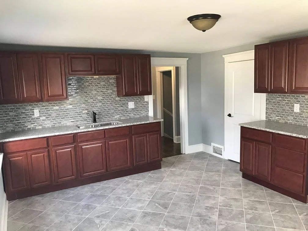 Kitchen with gray tiled floor, dark red cabinets, gray tile backsplash, black faucet, white door, and gray walls.