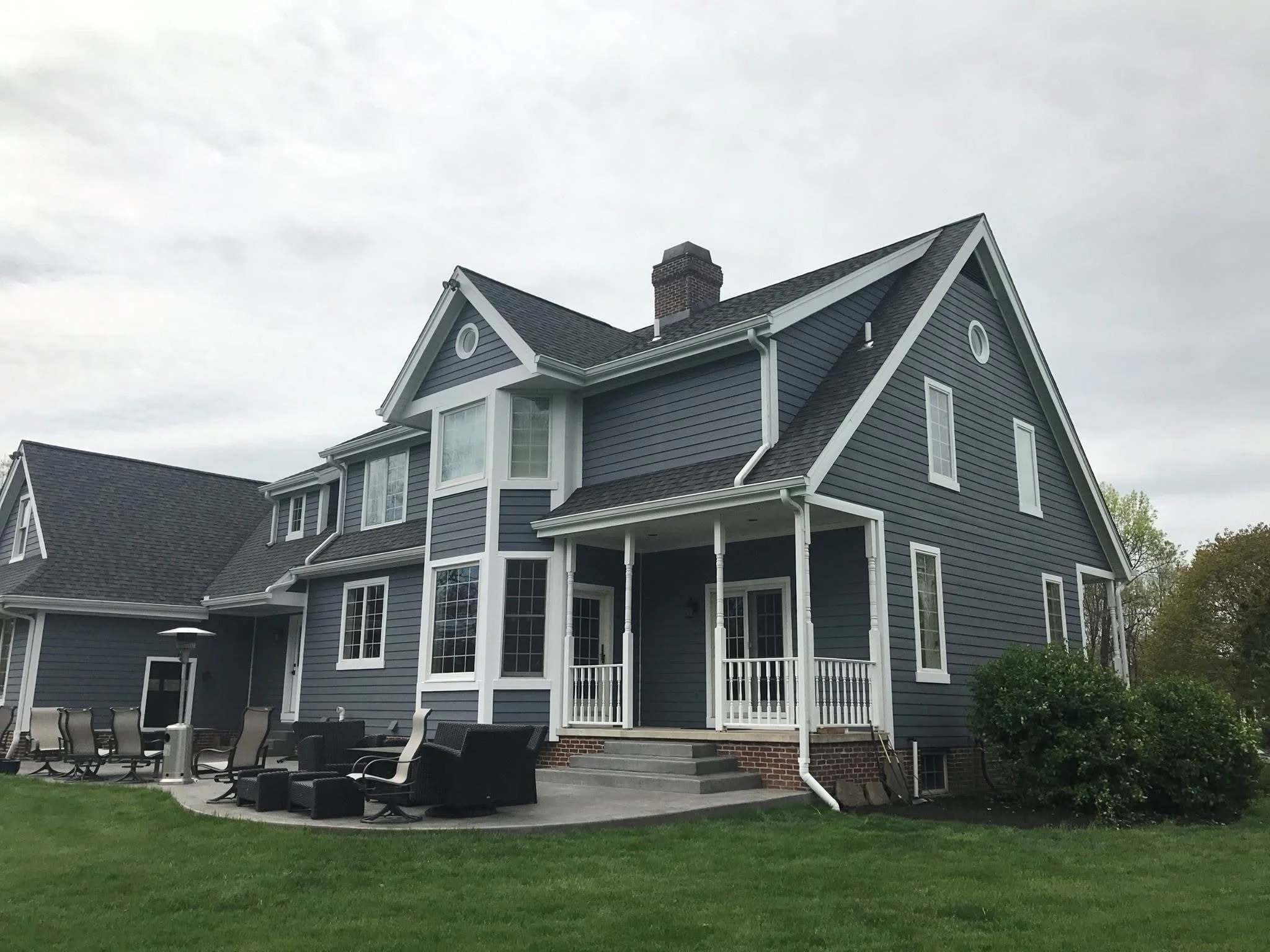 A large blue house with white trim, multiple windows, a porch, and a gray roof. The house has a brick chimney and a well-maintained green lawn with outdoor furniture and a patio area.