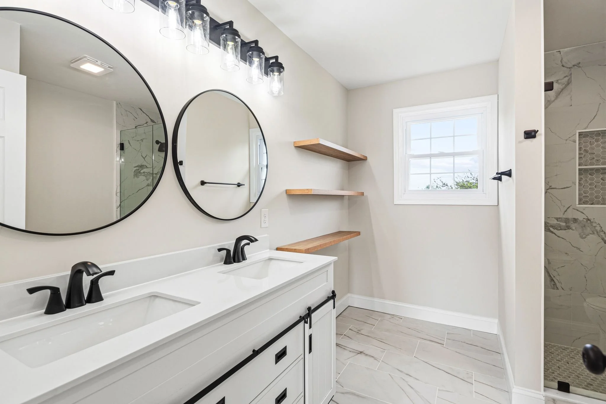 A modern bathroom with a double white vanity, black faucets, and large oval mirrors. Wooden open shelves are mounted on the wall, and there's a window letting in natural light. The bathroom has light-colored walls and marble-patterned tiles on the floor and shower wall.