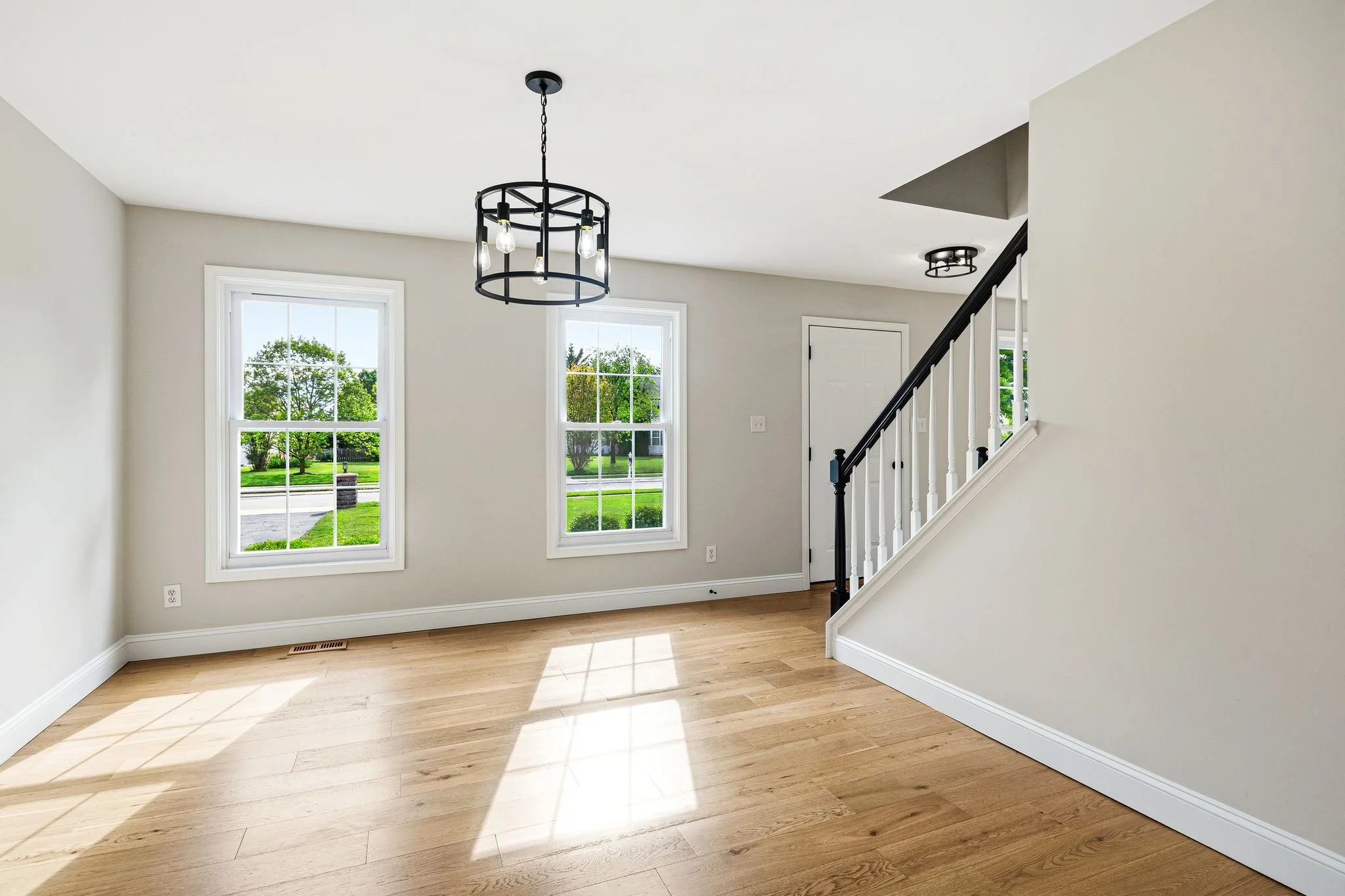 Empty living room with two large windows, a staircase with black and white railing, LVP floors, and two ceiling light fixtures.