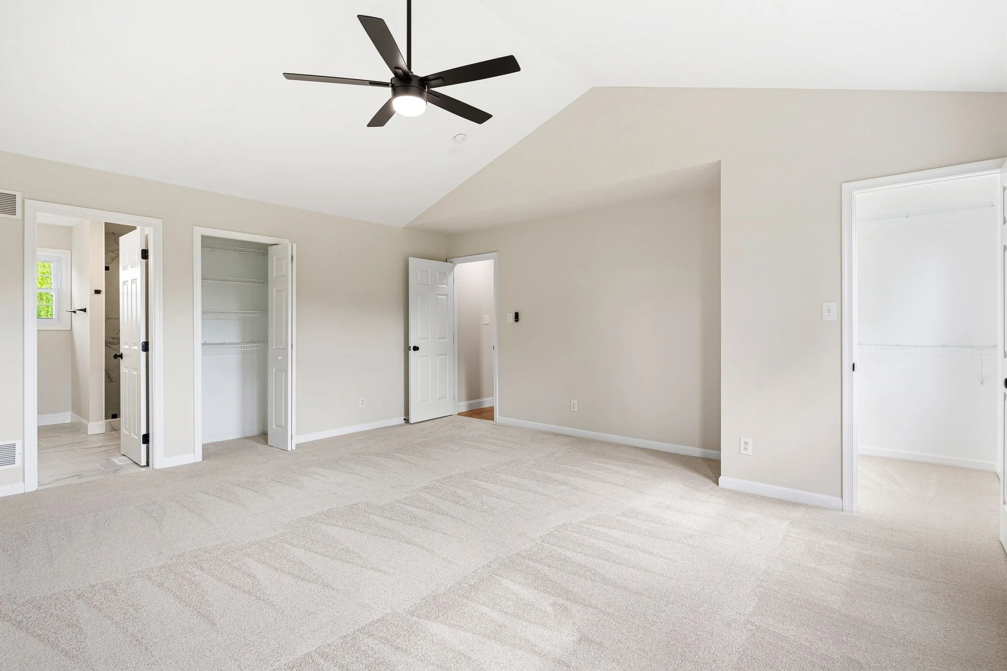 Empty beige-carpeted room with white walls and multiple open doors leading to closets and other rooms, ceiling fan with black blades and light, and natural light coming through a window.