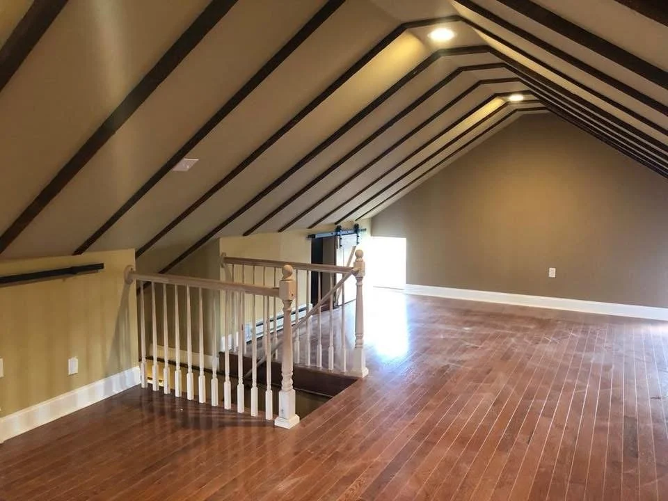 Empty attic bedroom with sloped ceiling, hardwood flooring, beige walls, and a small staircase with a white banister leading down to a door.
