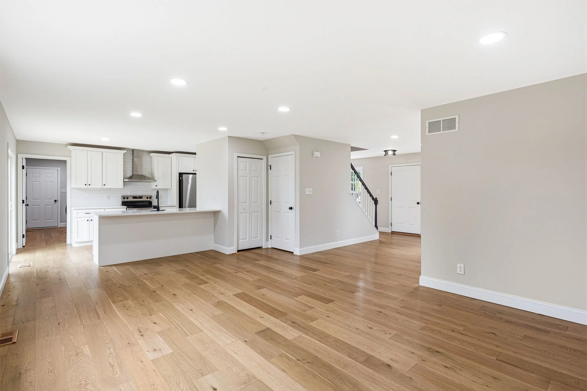 Open-concept living space with LVP floors, white walls, and a ceiling with multiple recessed lights. Kitchen area visible in the background with white cabinets, a stainless steel oven, and a black faucet.