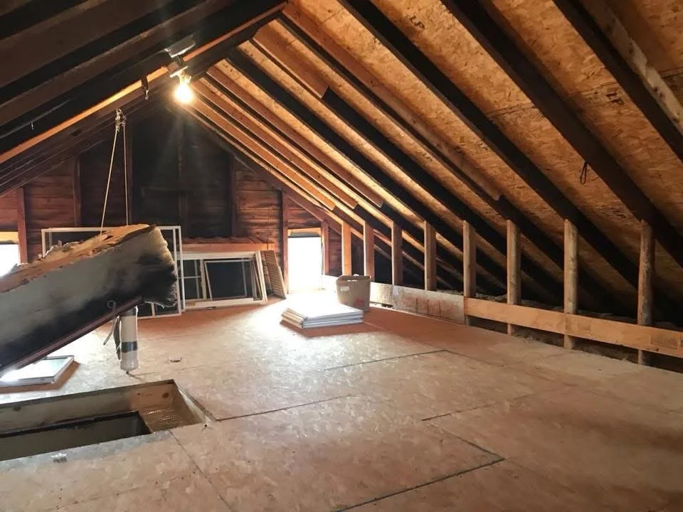 Attic under construction with exposed wooden framing, plywood flooring, and construction materials including a window frame, and insulation.