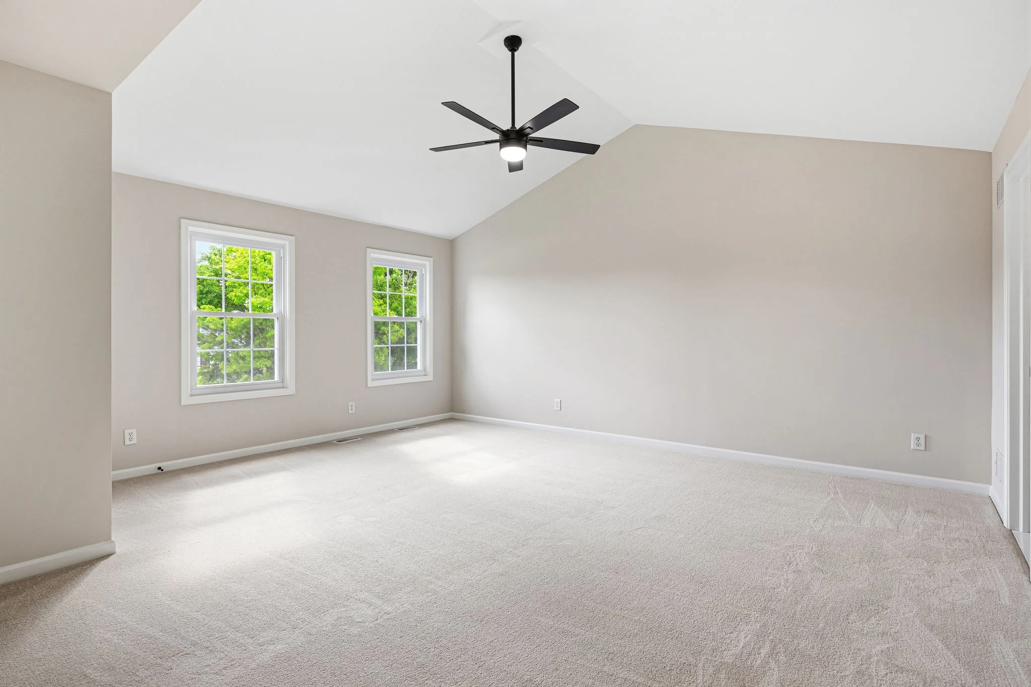 A bright empty room with beige walls, white trim, two windows with green trees outside, a ceiling fan with lights, and light-colored carpet.