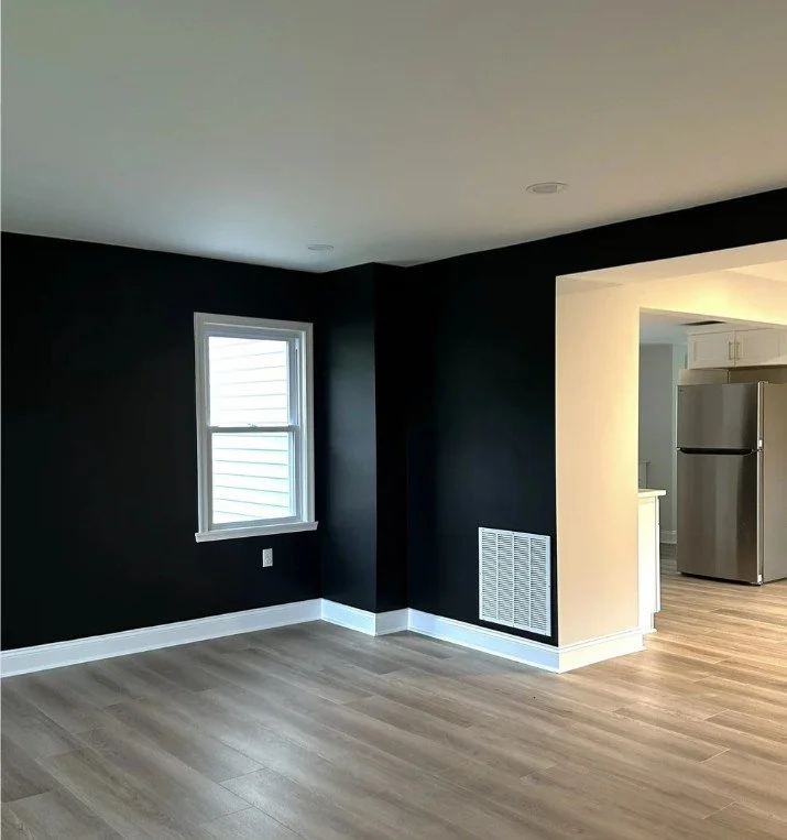 Empty room with dark walls, light wood flooring, a window, white baseboards, and an open doorway leading to a kitchen with stainless steel refrigerator.