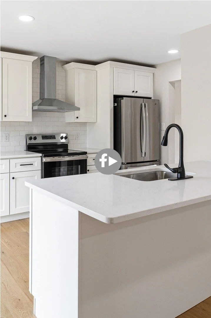 Modern white kitchen with stainless steel appliances, including a refrigerator, oven, and range hood. A white quartz countertop island with a black faucet and sink. Light wood flooring and recessed ceiling lights.