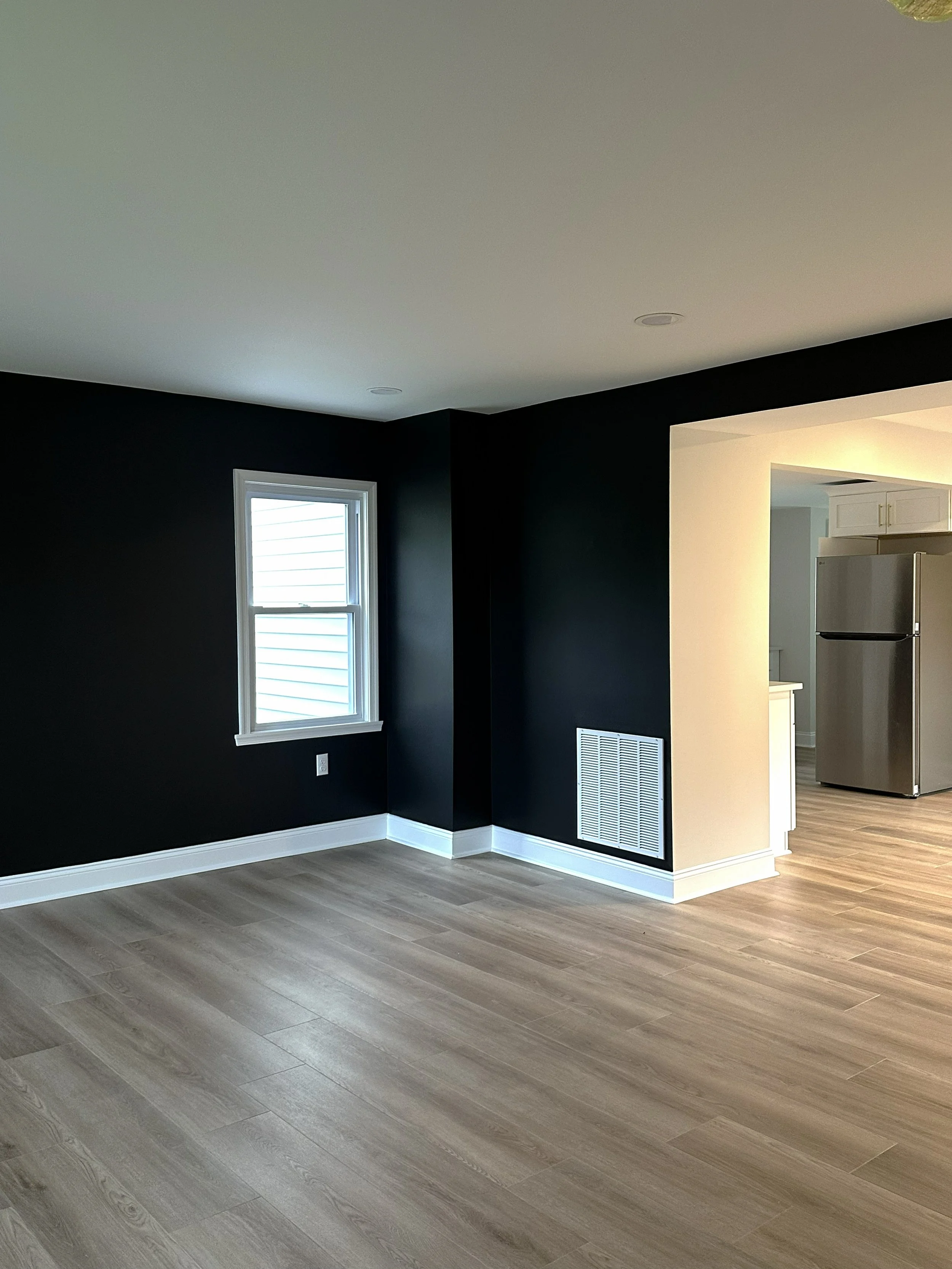An interior room with LVP flooring, black accent wall, white window frame, and a partial view of a kitchen with a stainless steel refrigerator.