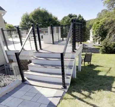 Outdoor staircase with black metal railings leading to a raised deck in a backyard with grass, trees, and outdoor furniture.