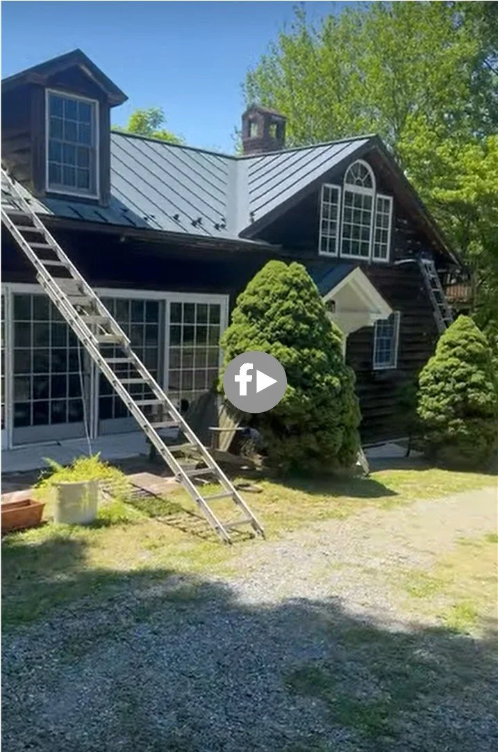A two-story dark wooden house with multiple large windows, a steep metal roof, and two ladders leaning against it, surrounded by green trees and a grassy area.