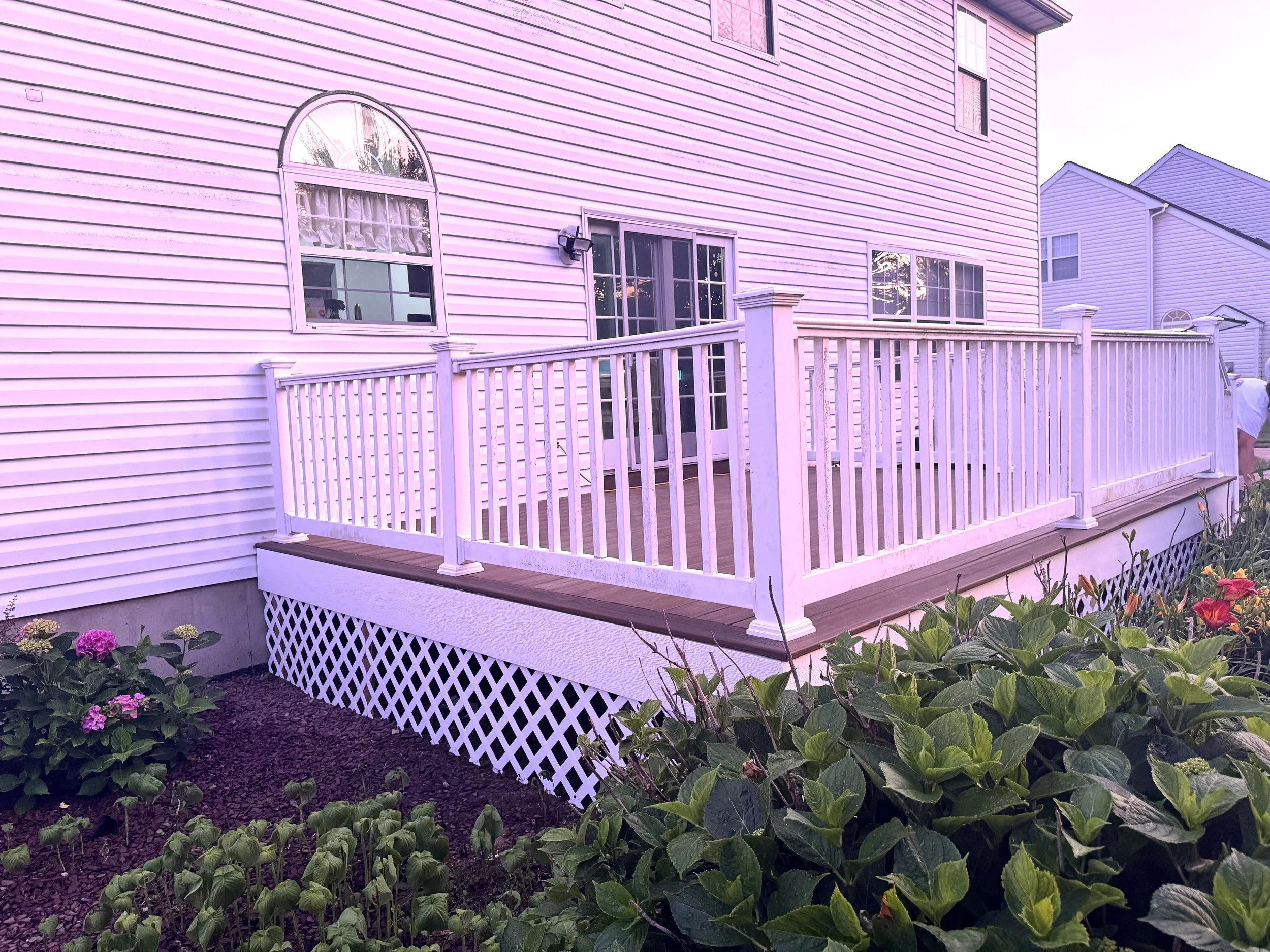 A house with pink siding and a wooden deck with white railing, surrounded by green plants and flowers.
